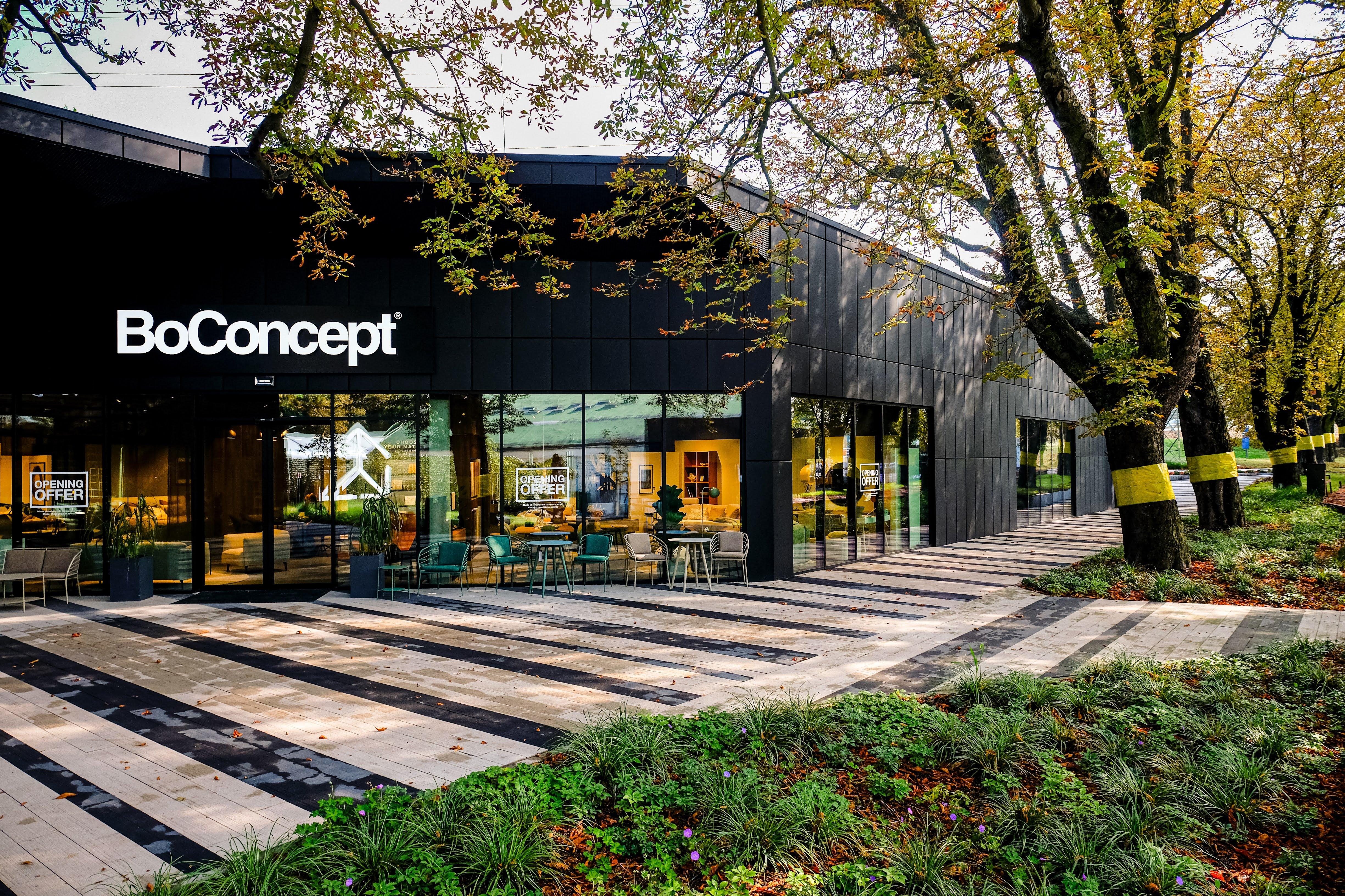 Fachada de tienda BoConcept con sillas y mesas exteriores. Árboles alineados a la derecha.