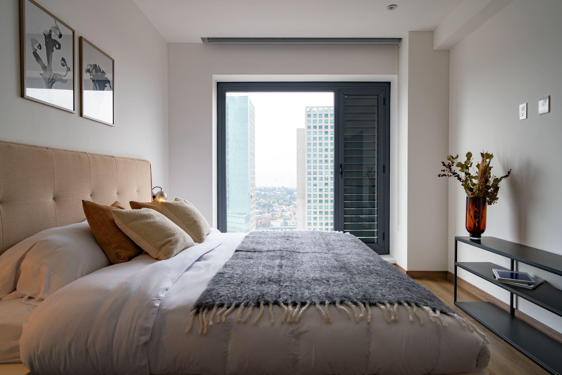 Modern bedroom with plush bedding, city view window, and minimalist decor.