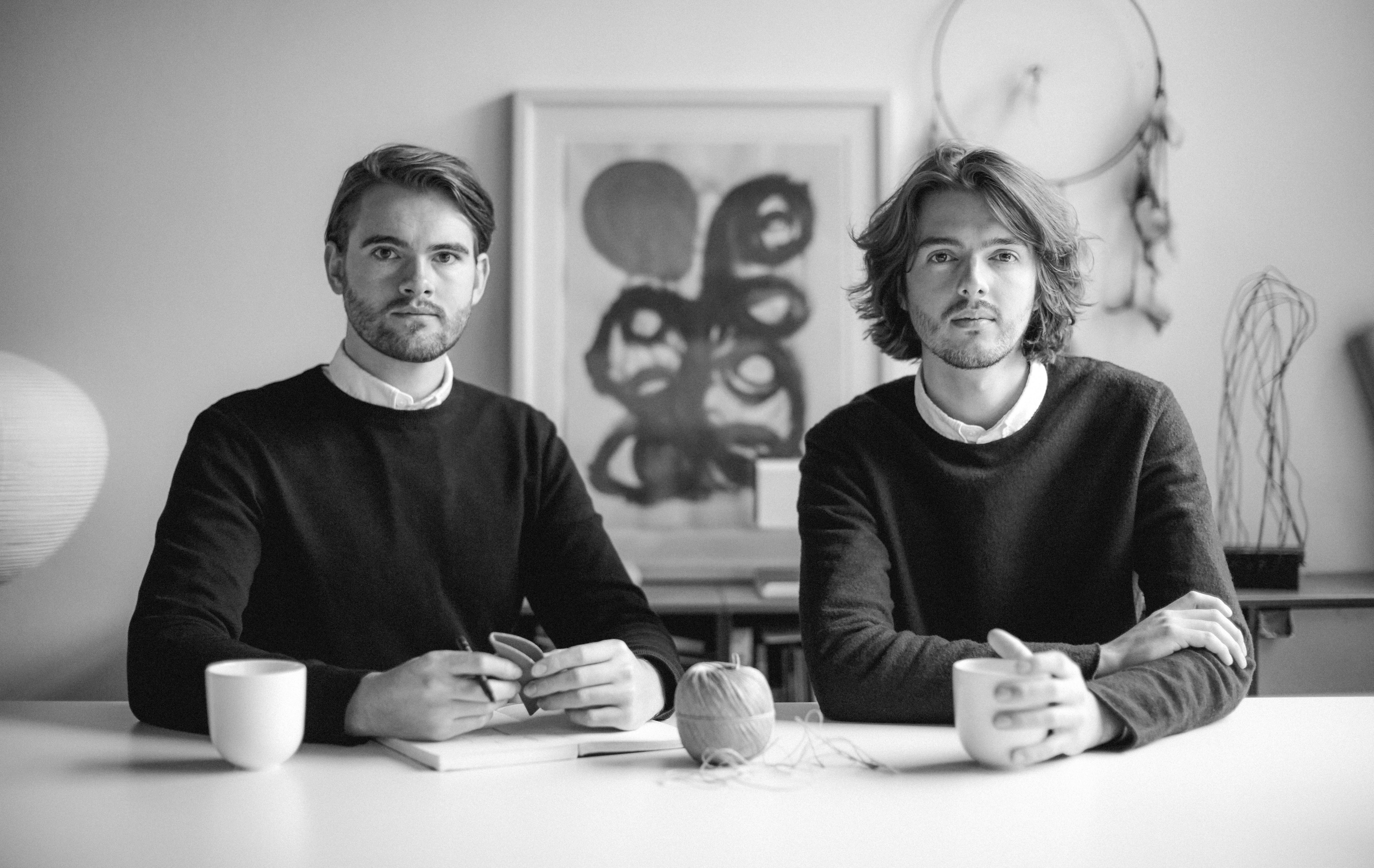 Two people sit at a table with notebooks, mugs, and a ball of yarn. Abstract art in the background.