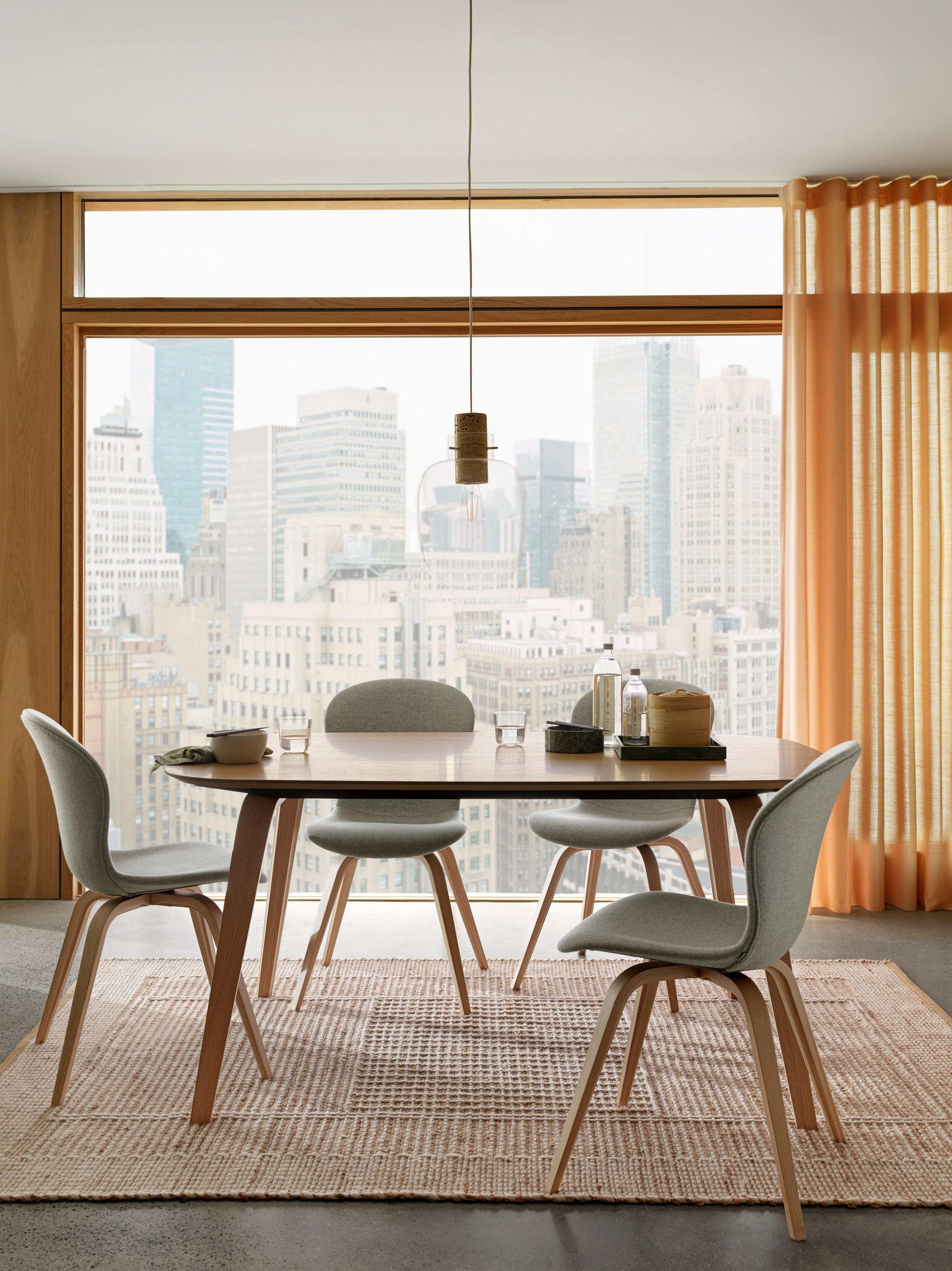 Dining area with table, chairs, and pendant light, city view through large window, on a woven rug.