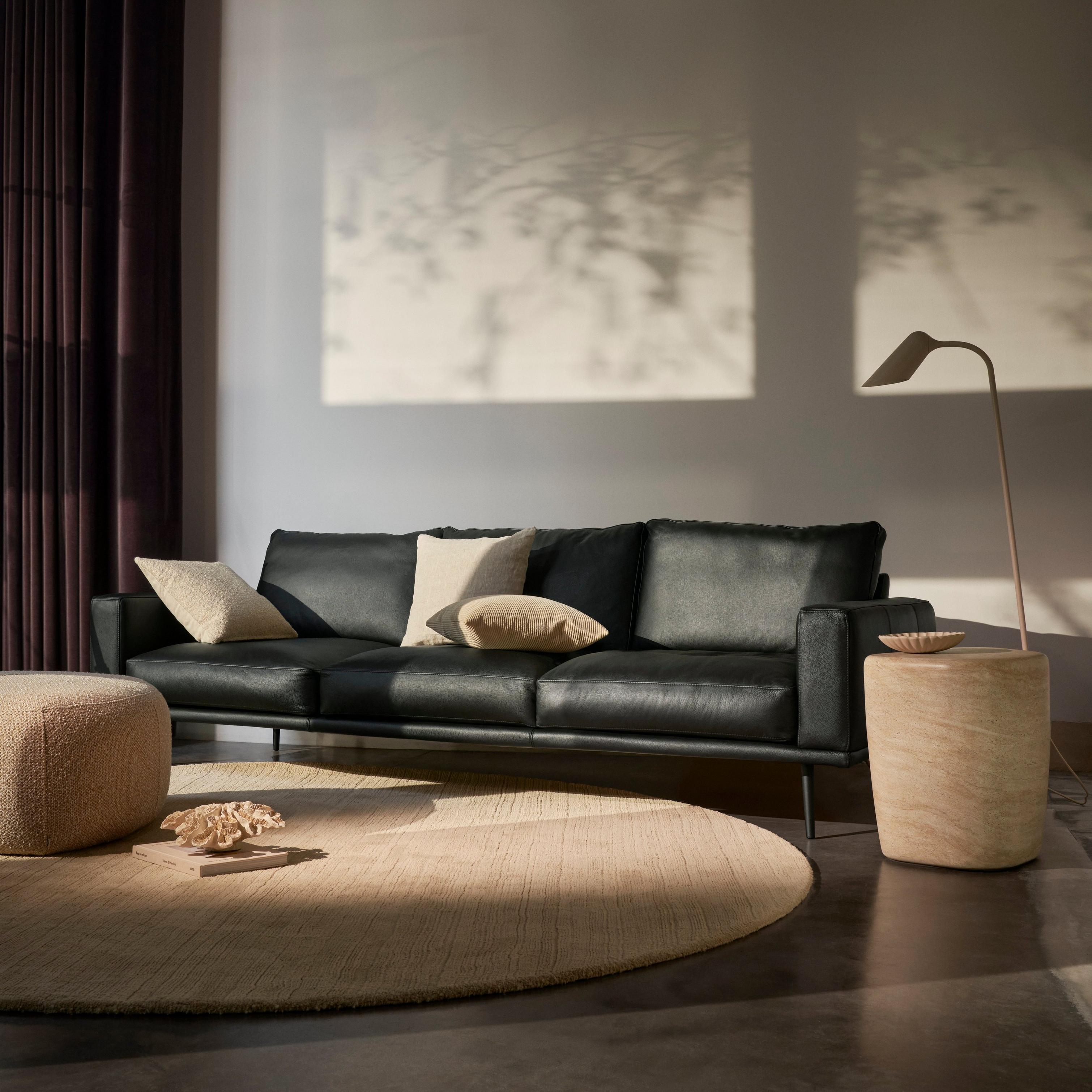 Modern living room with a black leather Carlton sofa, beige pillows, round rug, ottoman, side table, floor lamp, and dark curtains.