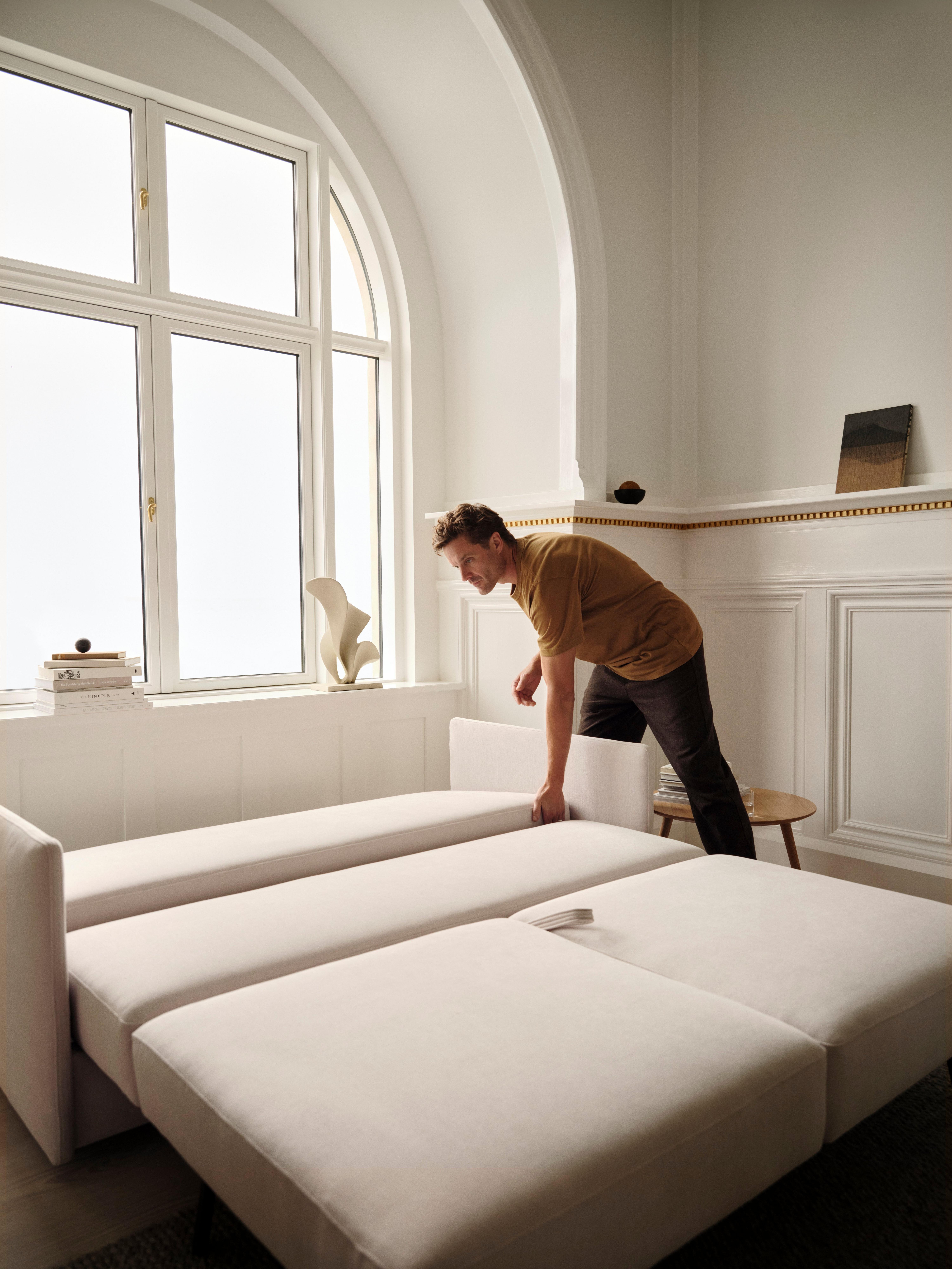 Man adjusting a convertible sofa bed in a bright room with large windows.