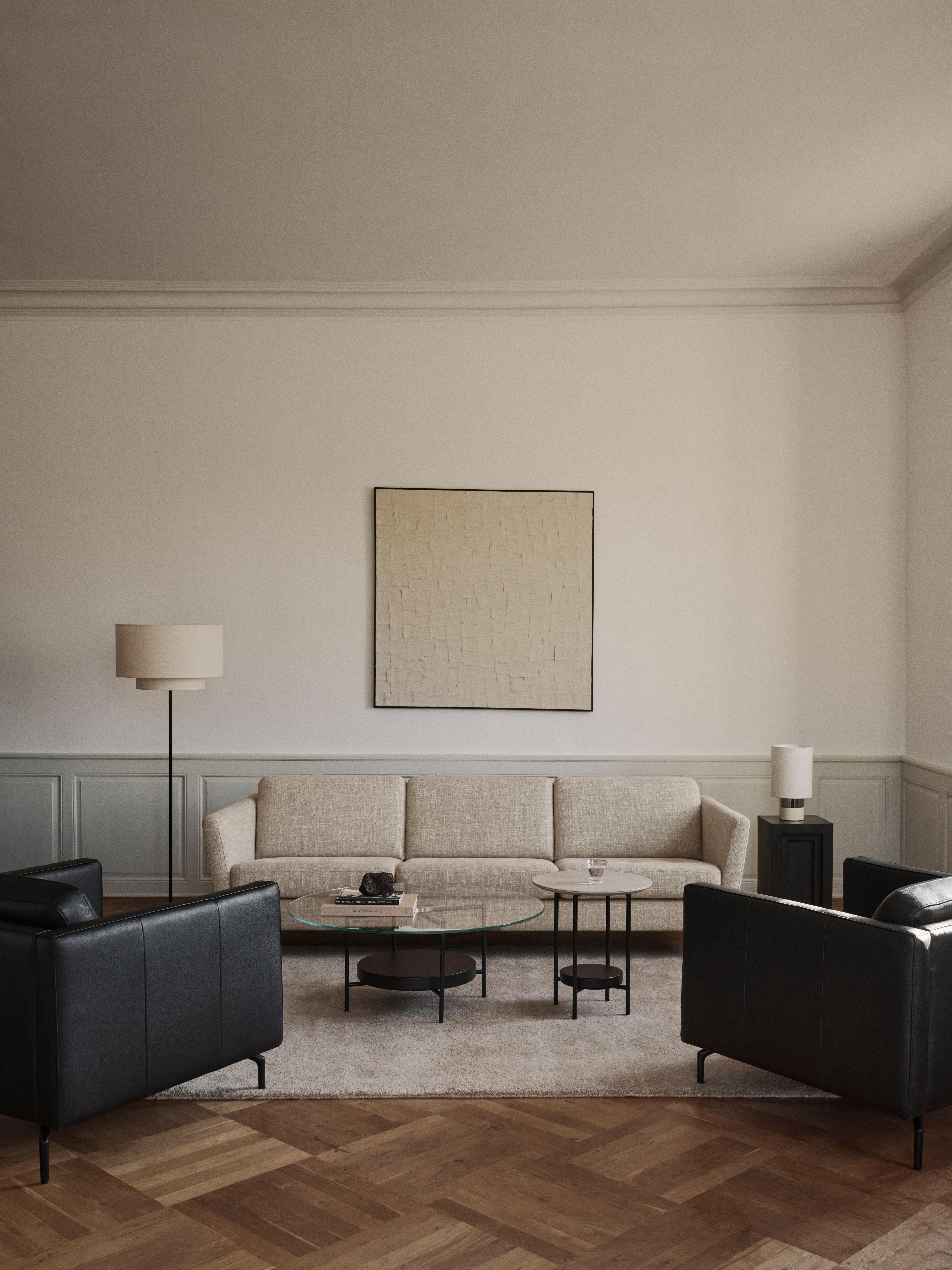 Salon avec canapé beige, deux fauteuils noirs, table basse en verre et lampadaire.