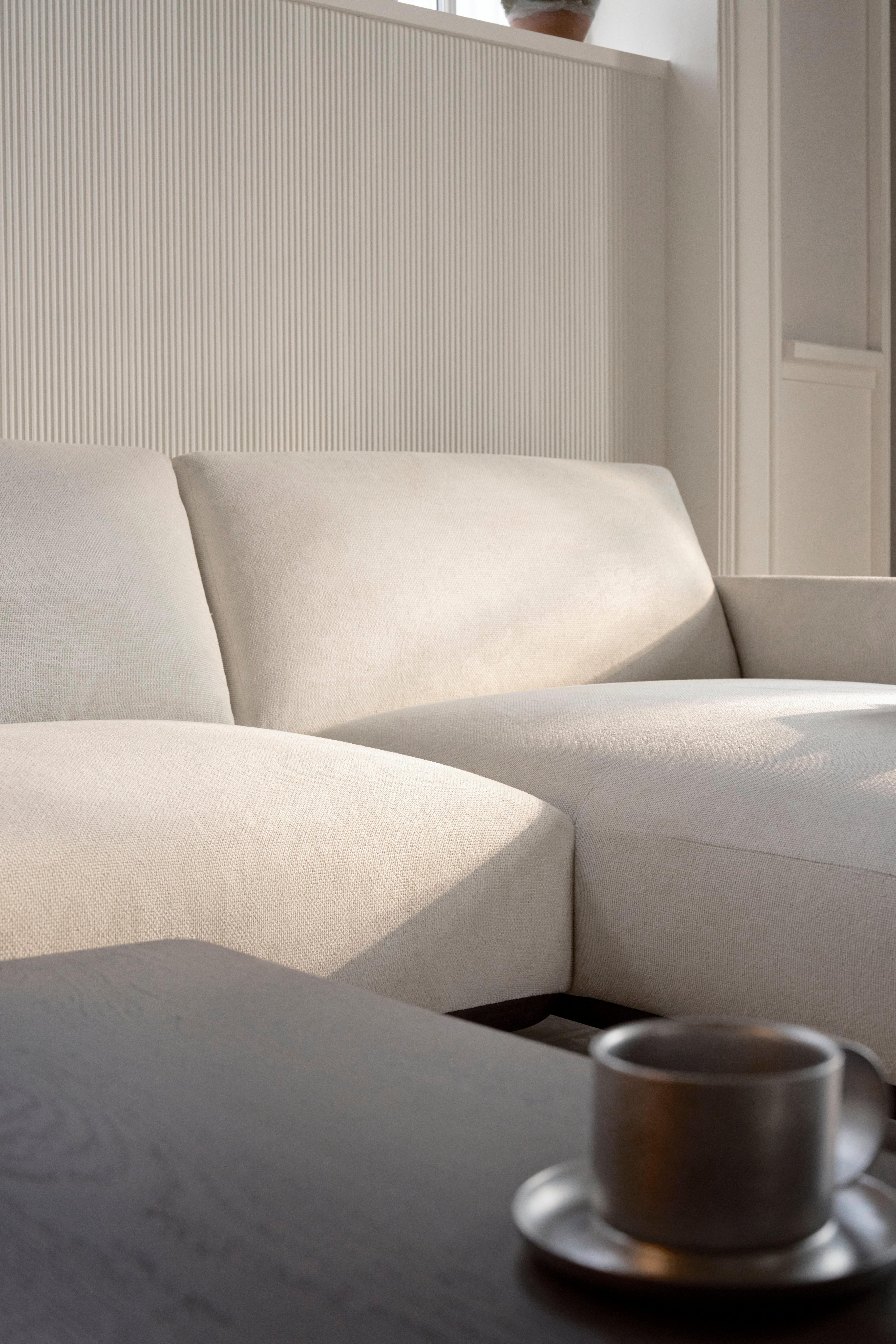 Jasnobeżowa narożna sofa z pochylonymi oparciami i podłokietnikami, ustawiona za ciemnym drewnianym stolikiem kawowym, na którym stoi metalowy kubek.