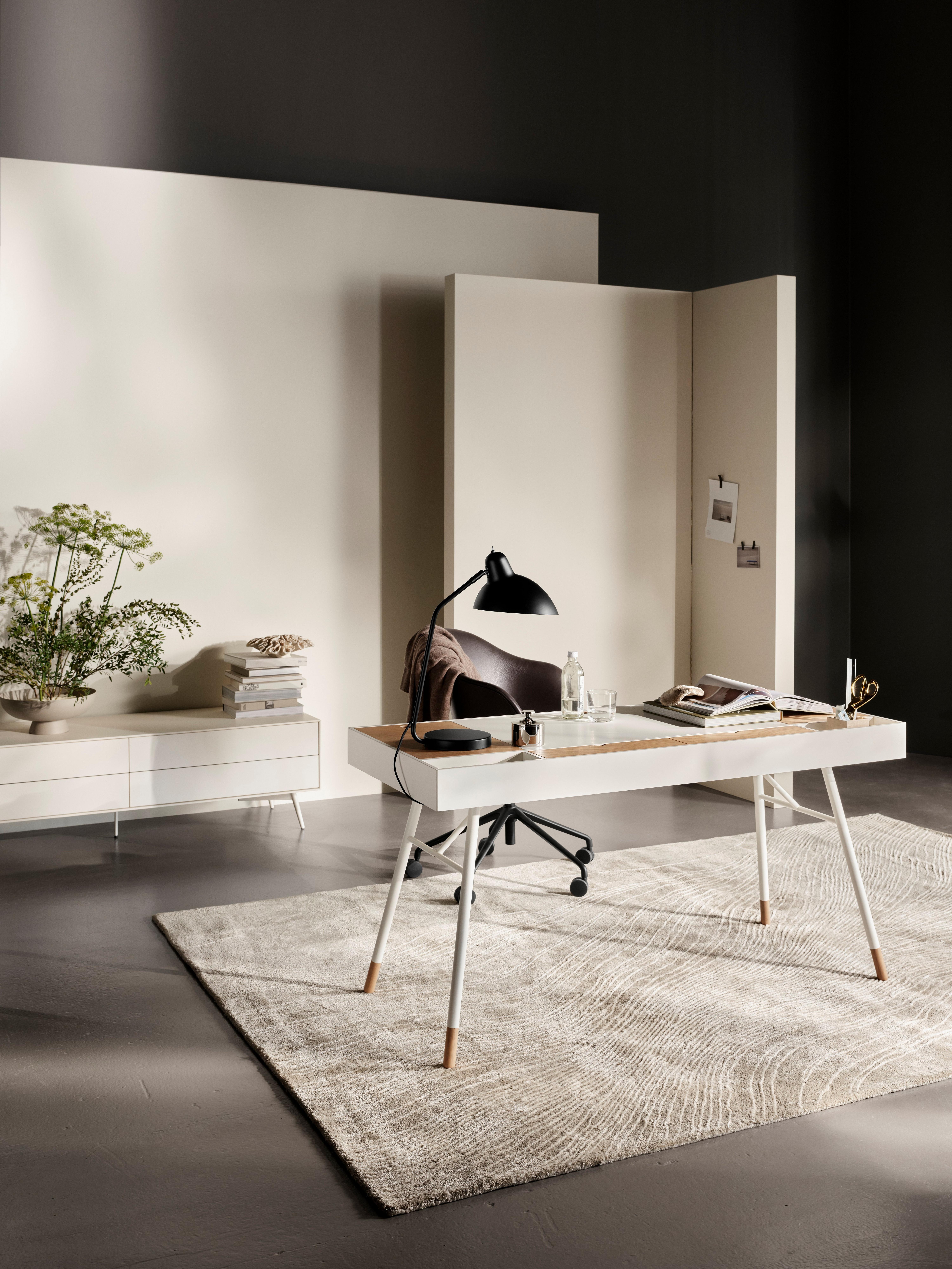 Modern office featuring a white lacquered Cupertino desk, white Fermo media unit, and a dark brown Adelaide office chair. A grey and white Tide rug anchors the space, with a Luna metal dish in warm grey on the desk. The matt black Demand table lamp illuminates the area, complemented by natural and warm grey Raja and Bali sculptures.