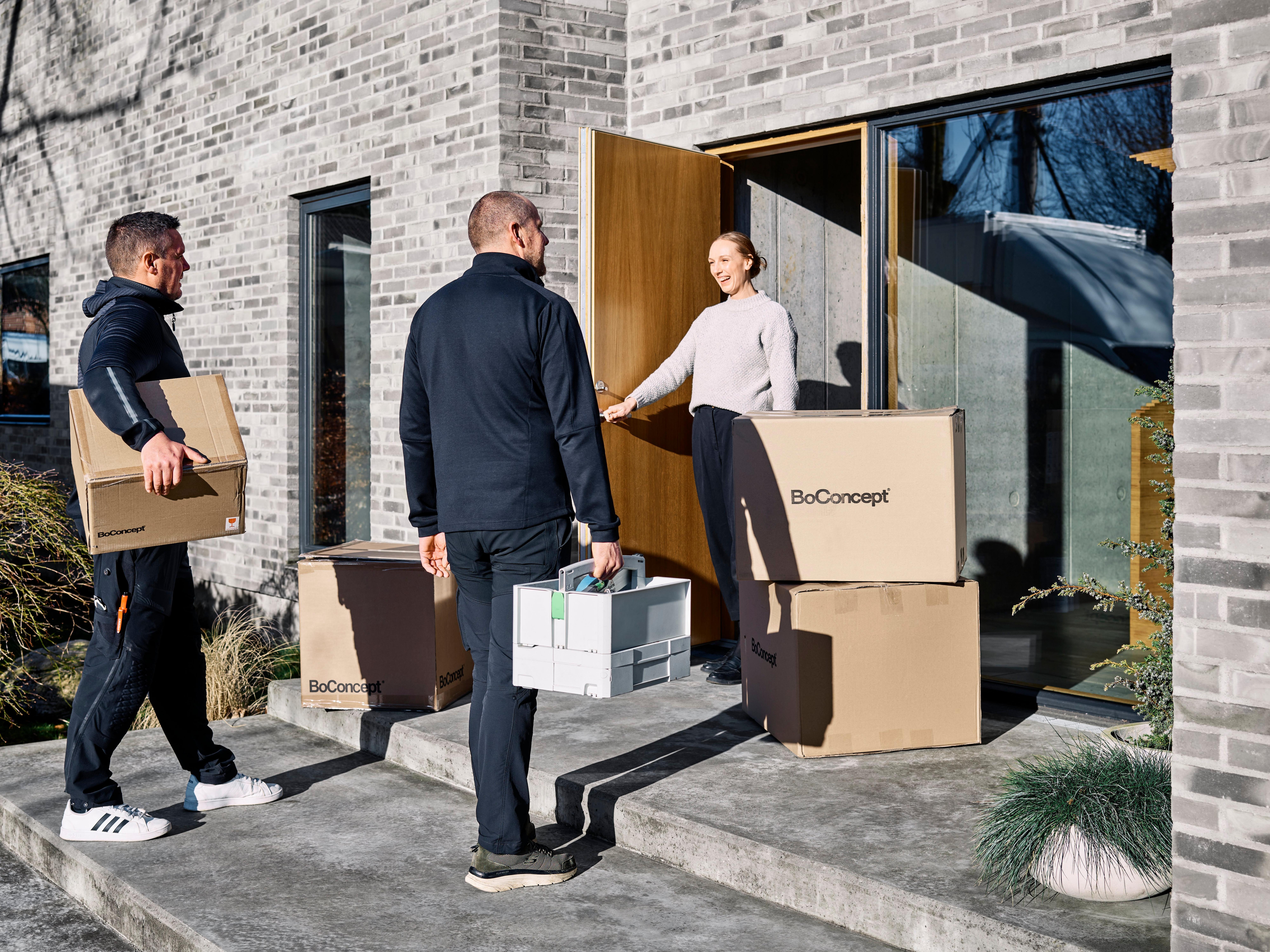 Two men deliver BoConcept boxes to a woman at the entrance of a modern grey-brick home with a wooden door.