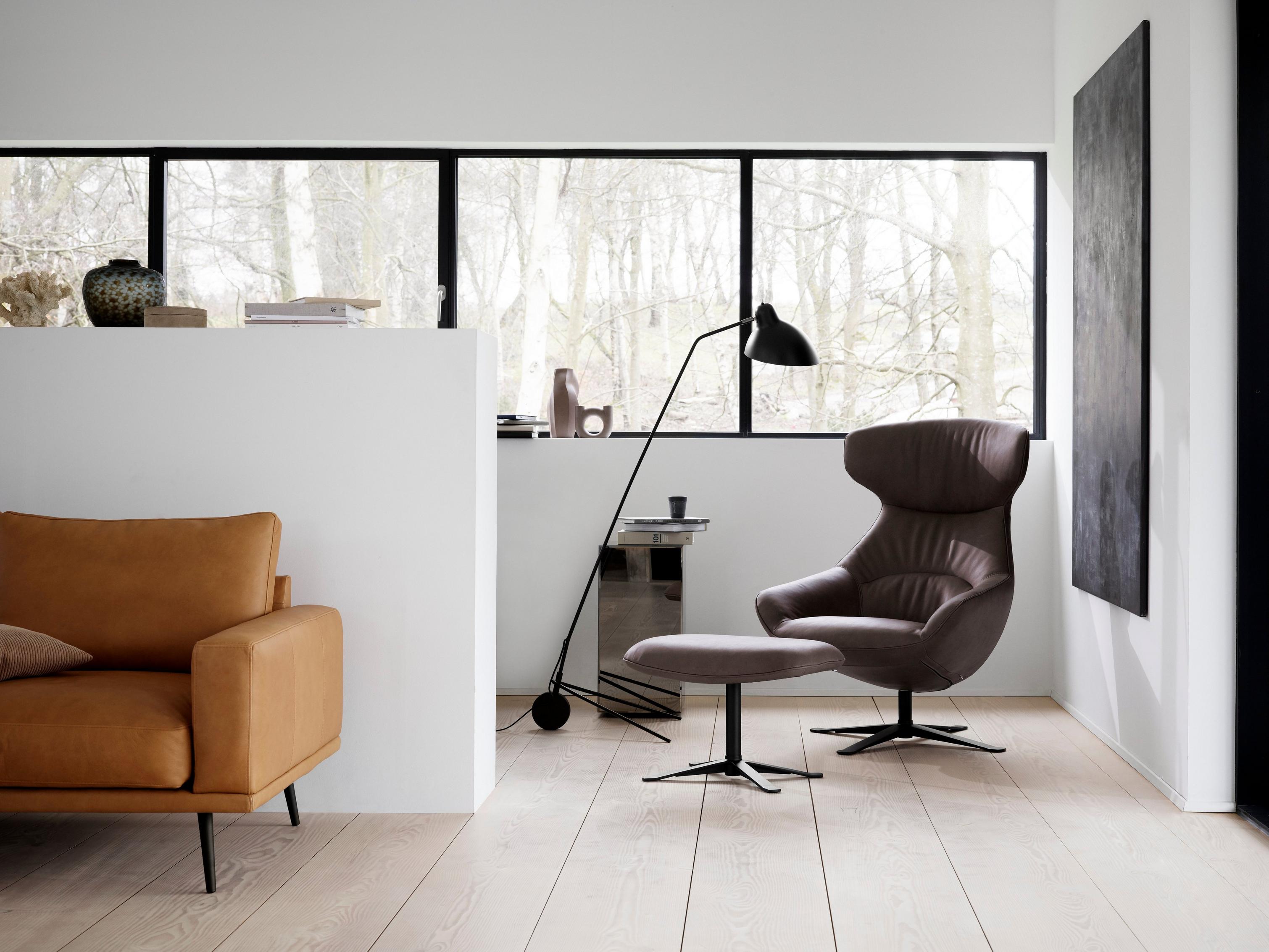 Modern reading corner with a tan armchair, dark lounge Porto chair and footrest, floor lamp, and forest view.