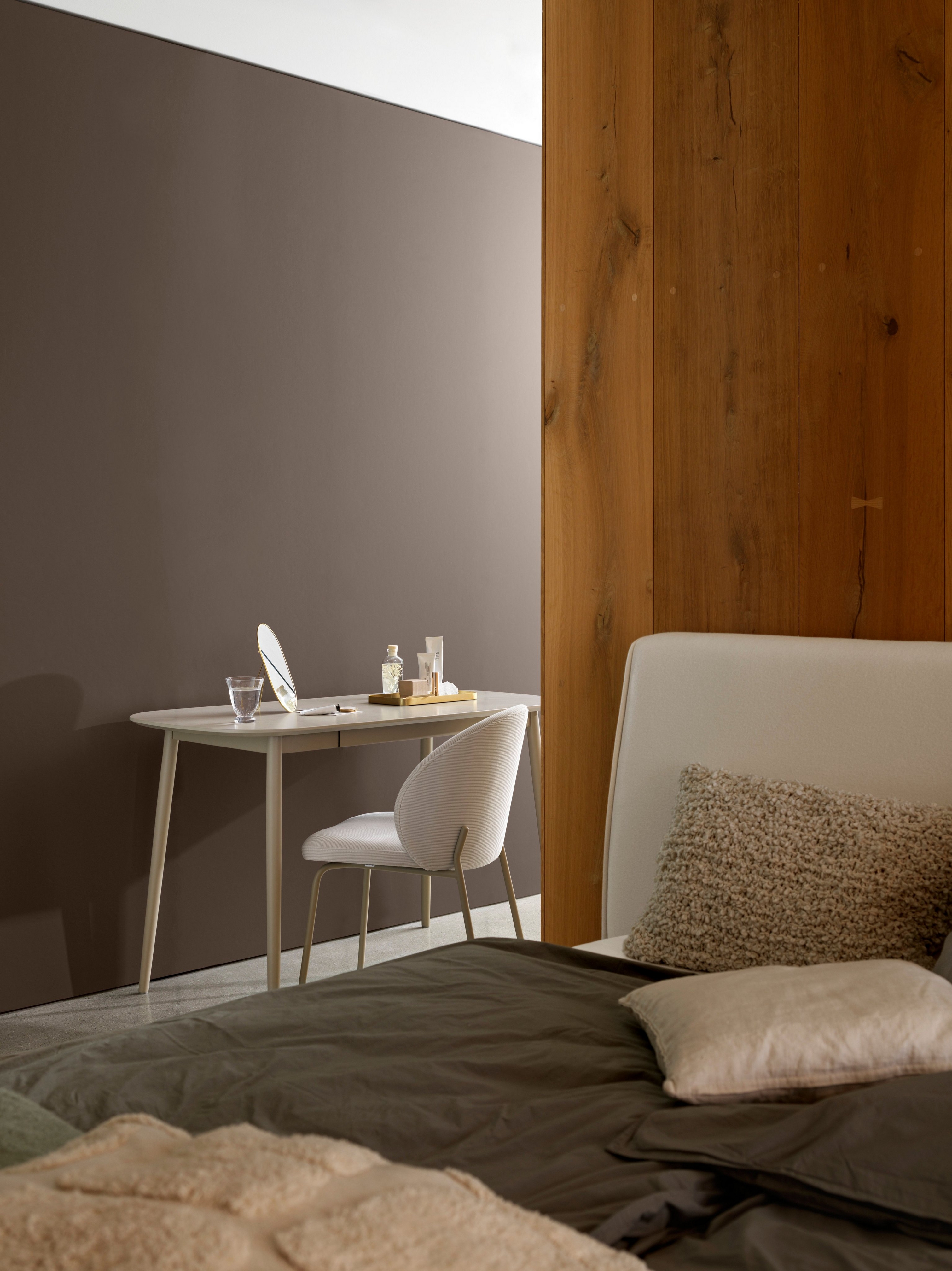 Roh spálne s jednoduchým stolom, stoličkou a útulnou posteľnou bielizňou pri drevenom paneli.