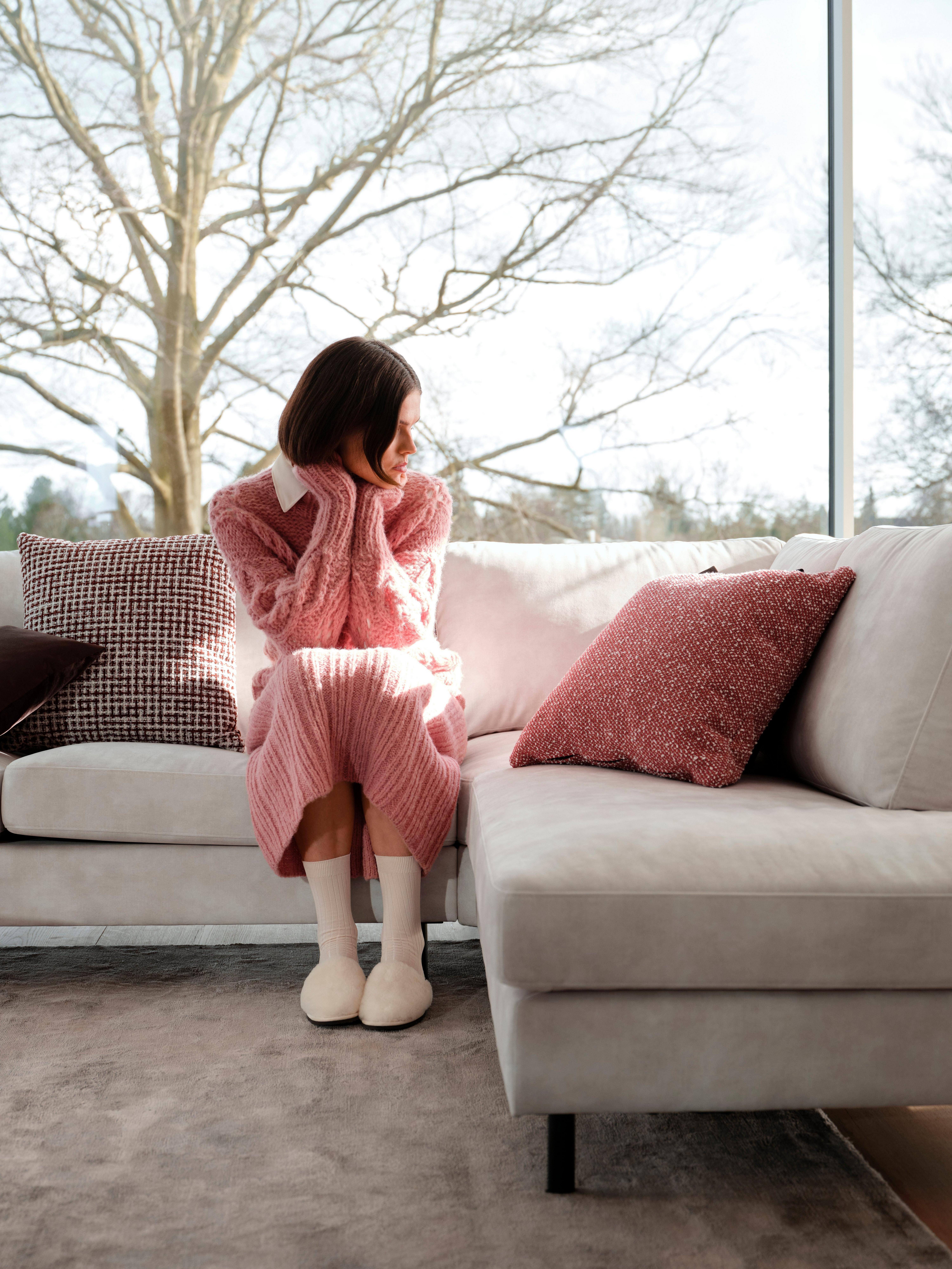 Person in a pink sweater and dress sitting on a Indivi corner sofa in beige Ravello fabric with Salamanca cushion in burgundy Tuscany fabric and red Avellino fabric and Velvet cushion in dark brown in a bright room with large windows.