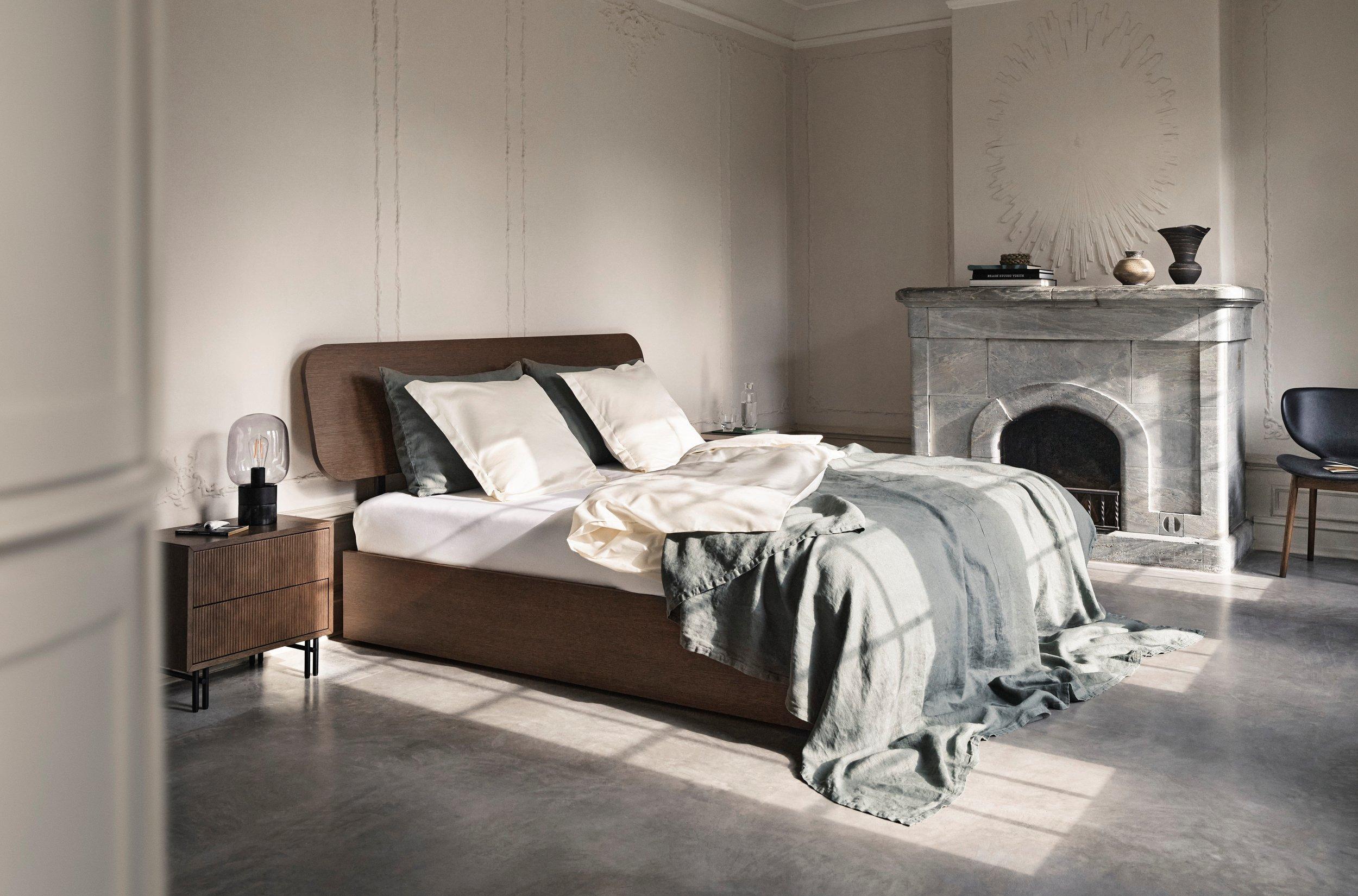 Bright bedroom with a brown upholstered bed, white and gray bedding, stone fireplace, and minimalist furniture in a classic interior.