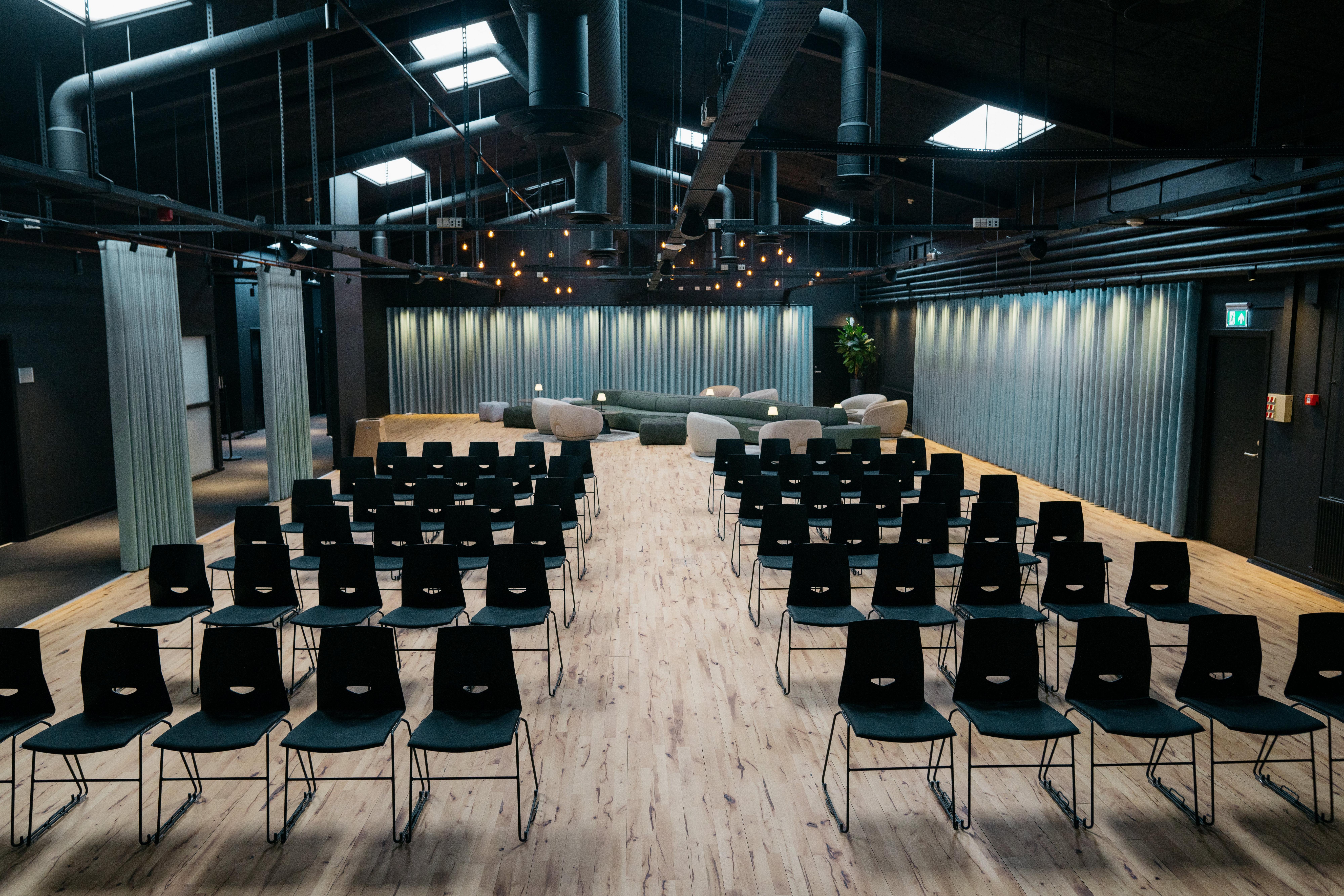 會議室有一排黑色椅子，面向採用綠色組合沙發、米色扶手椅和圓桌的休息室。木頭地板、藍色窗簾和工業用吊燈。