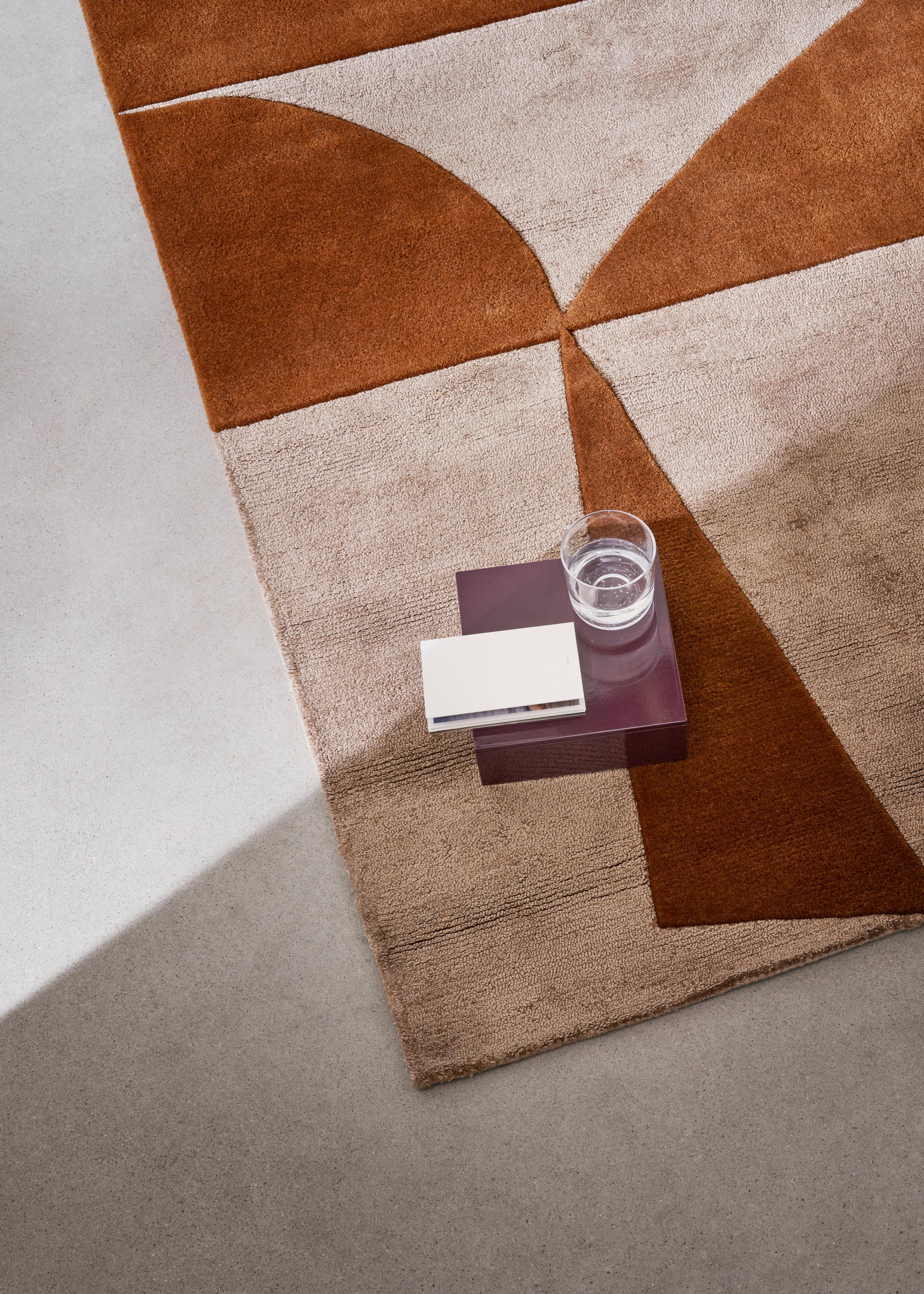 The Moments rug in beige and rusty orange with a geometric pattern is partially covered by a small purple side table holding a glass of water and a white book.