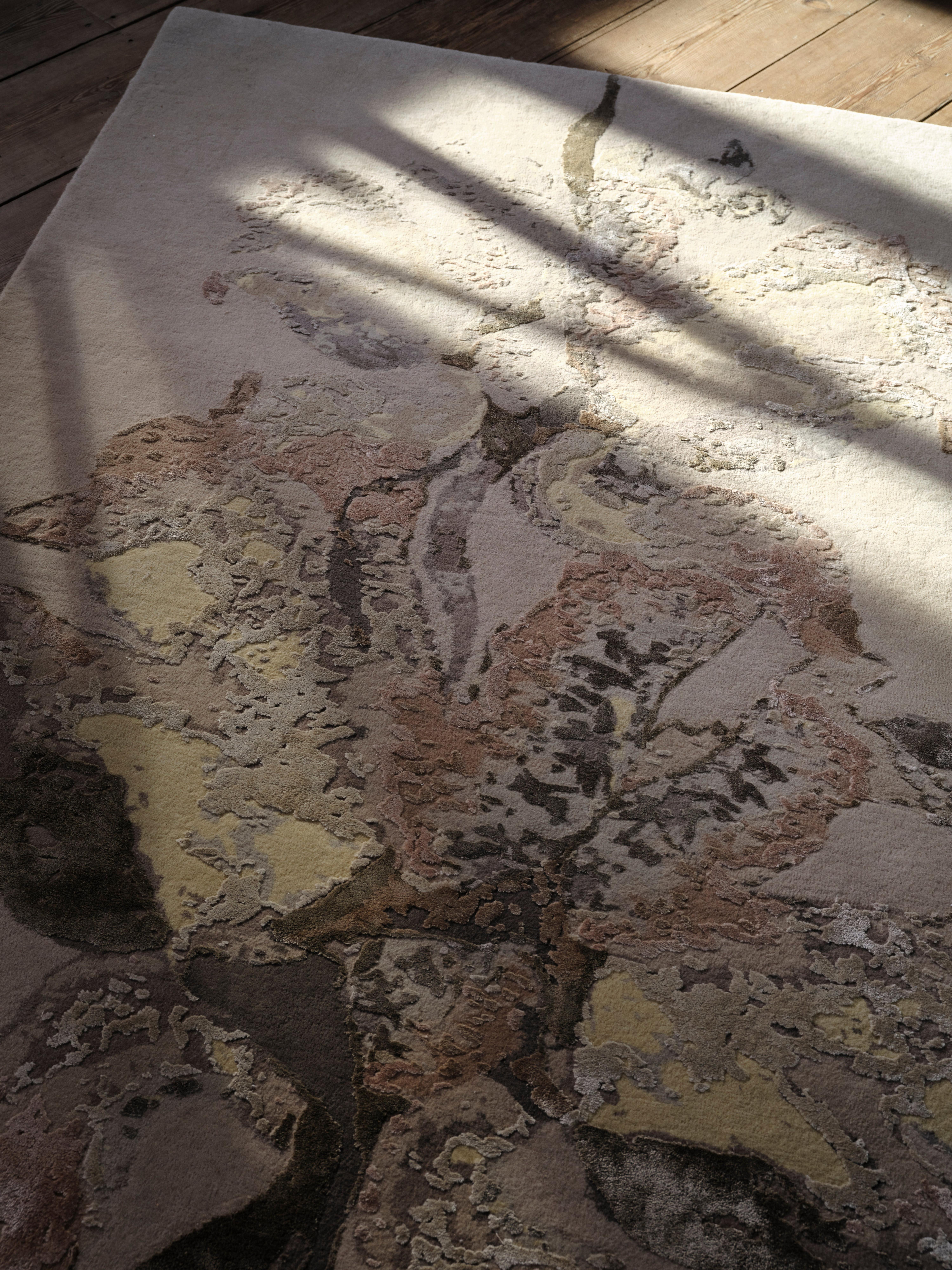 The Moments rug in beige and rusty orange with a geometric pattern is partially covered by a small purple side table holding a glass of water and a white book.
