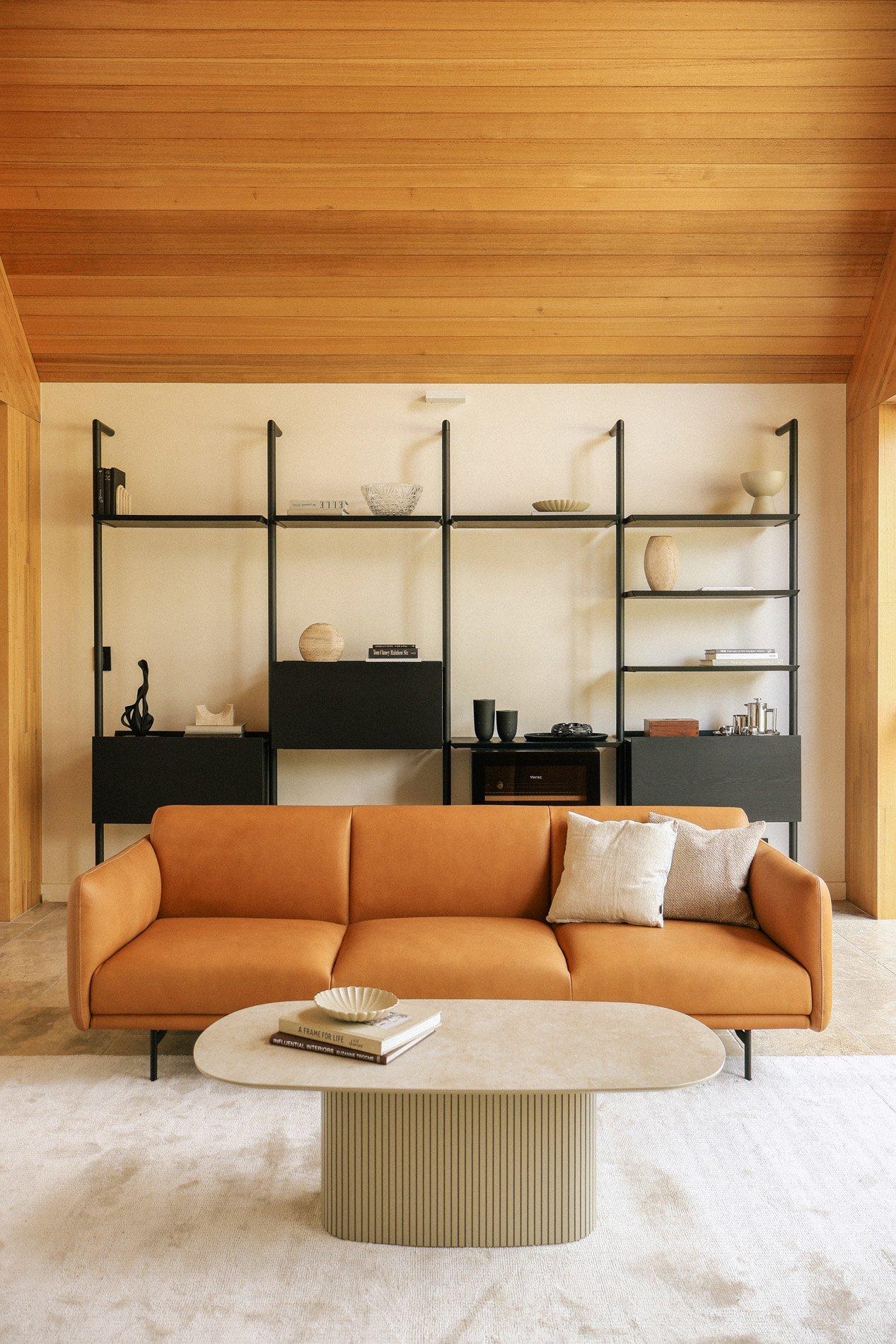 Tan three-seat sofa with cushions in front of black shelving unit and oval coffee table.