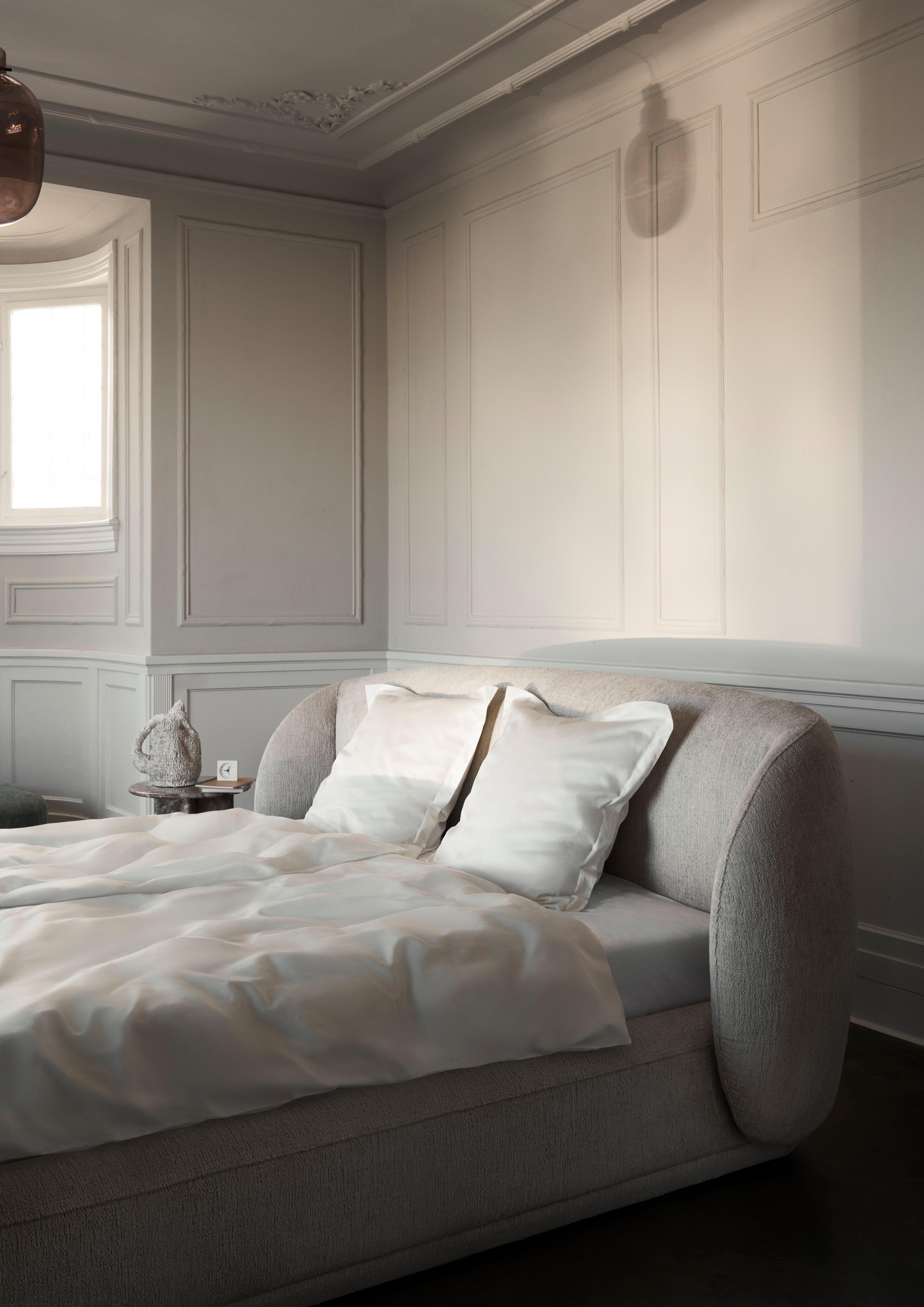 Elegant bedroom with a light gray upholstered bed, white bedding, and paneled walls, styled with soft lighting and minimal decor.