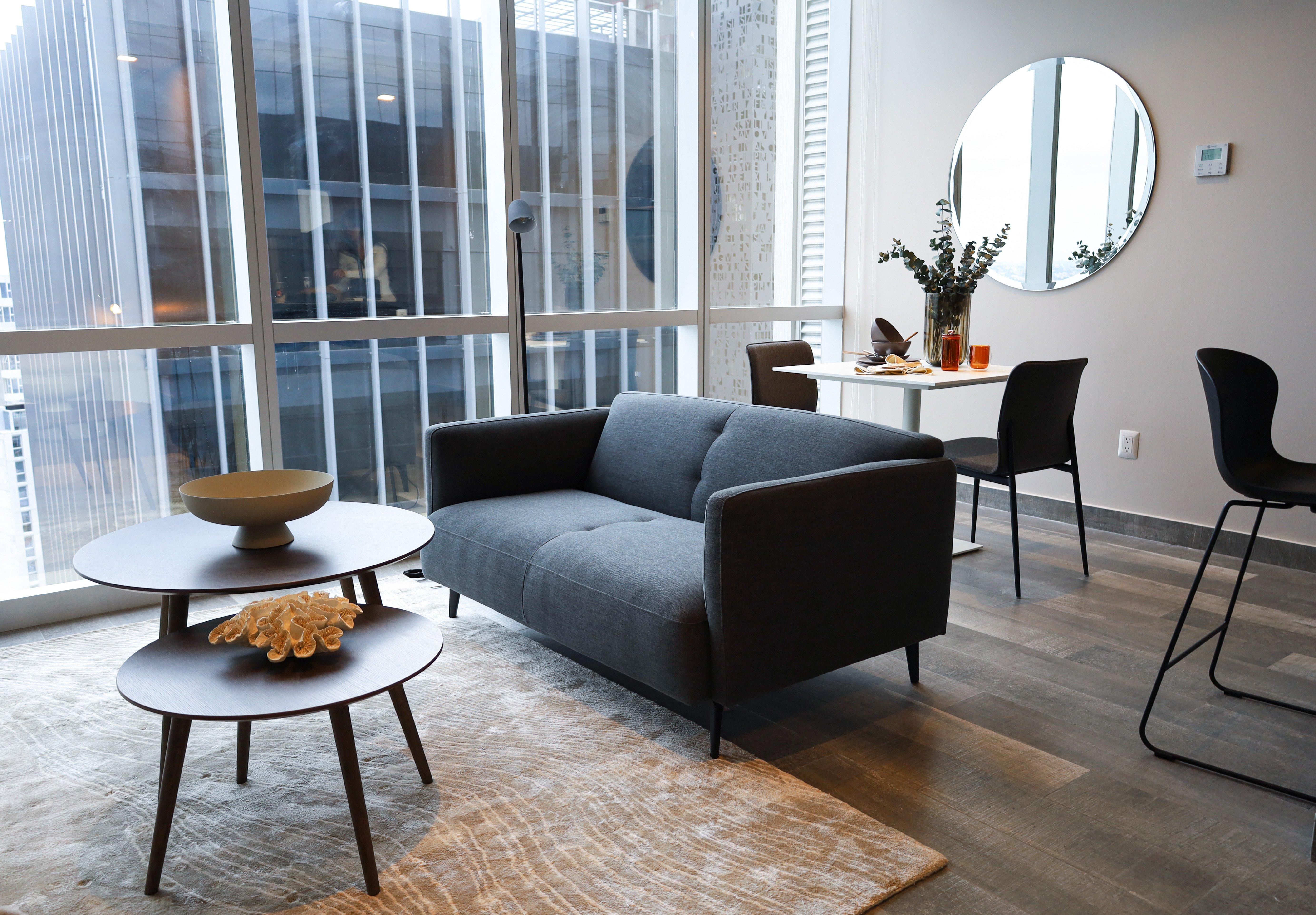 Modern living room with grey sofa, round tables, and a dining area by large windows. Mirror on wall.