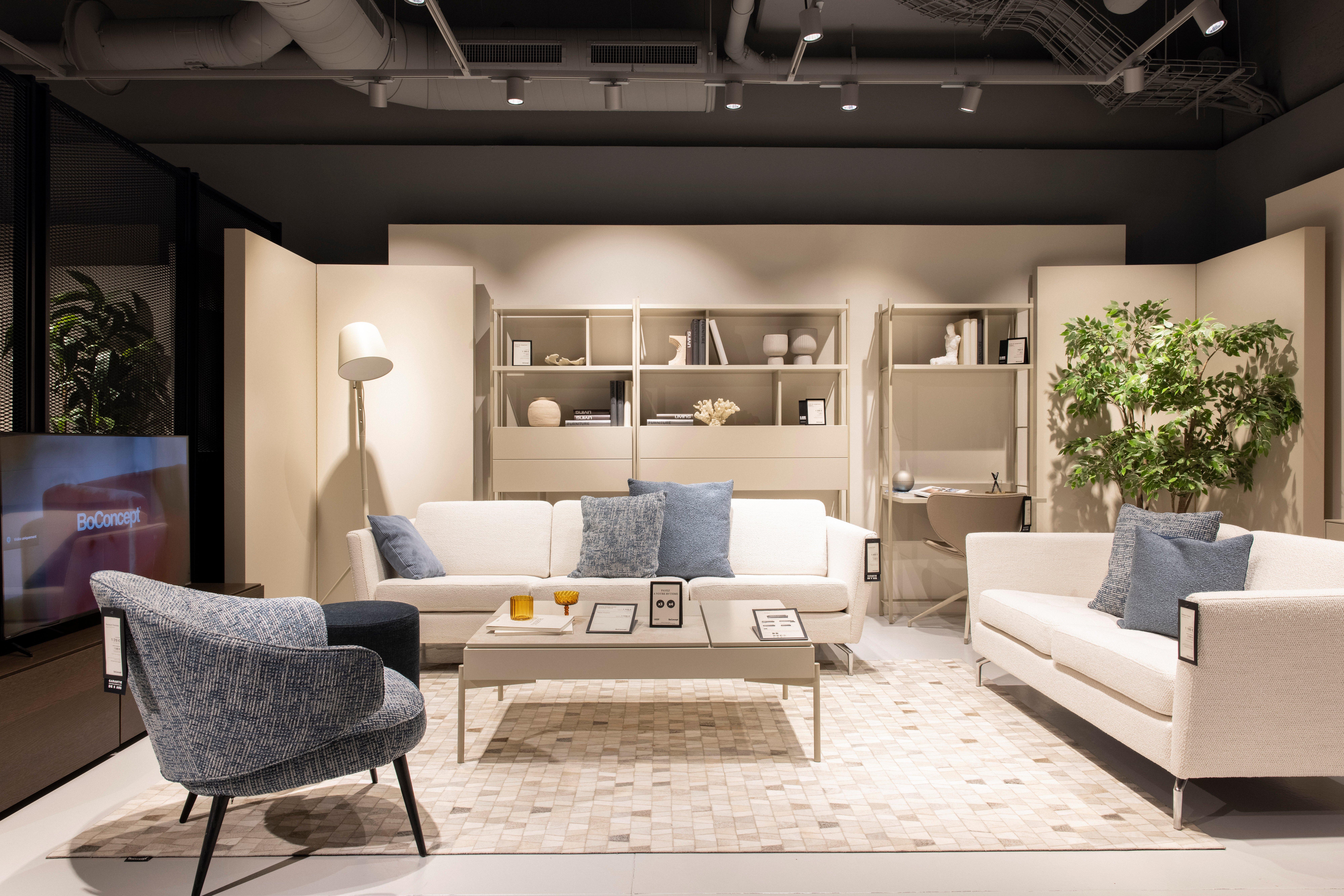 Moderne stue med to hvite sofaer, blå puter, et bord og en grønn plante. BoConcept-logo på TV.