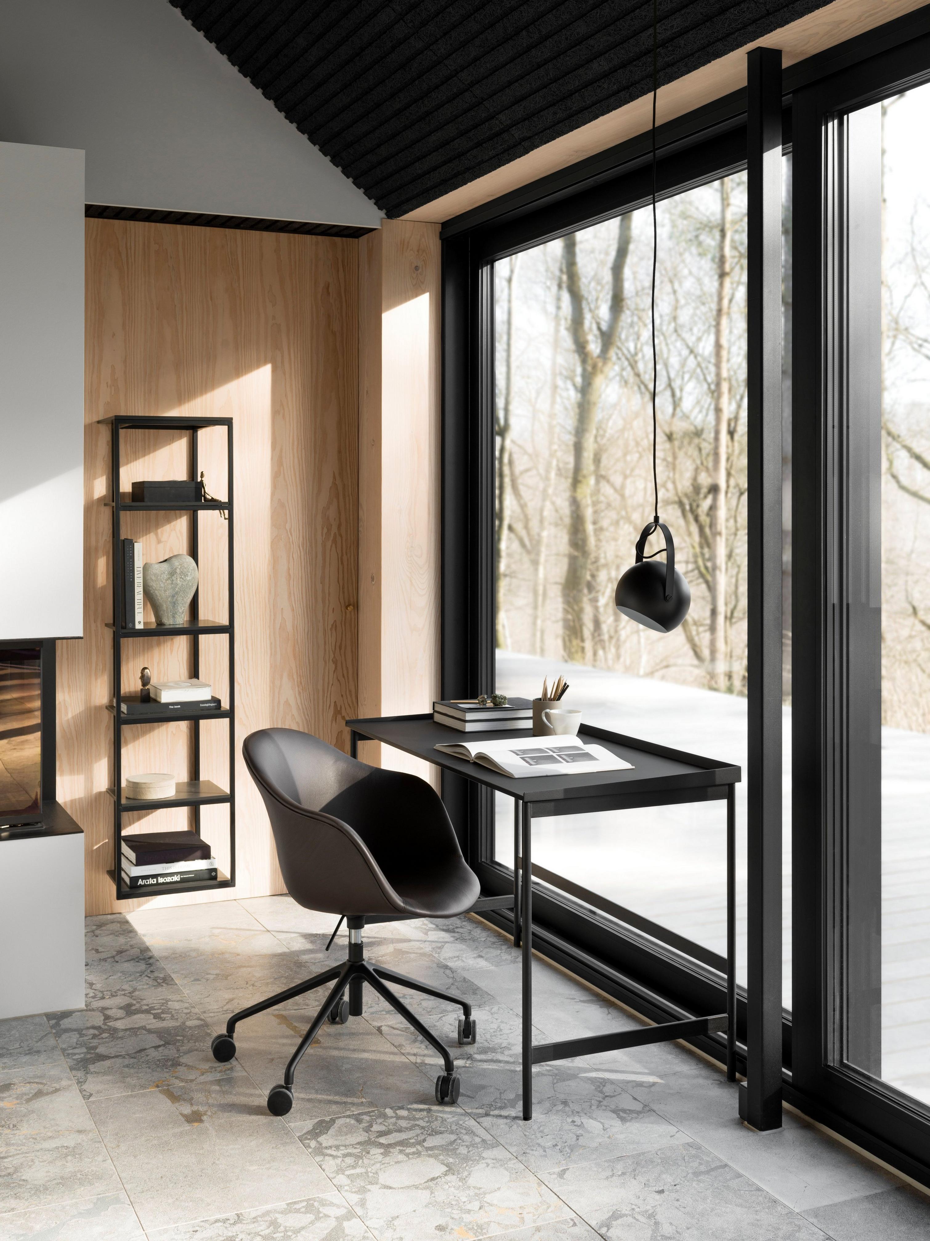 Modern office with black chair, desk, pendant light, and bookshelf by a large window with a forest view.