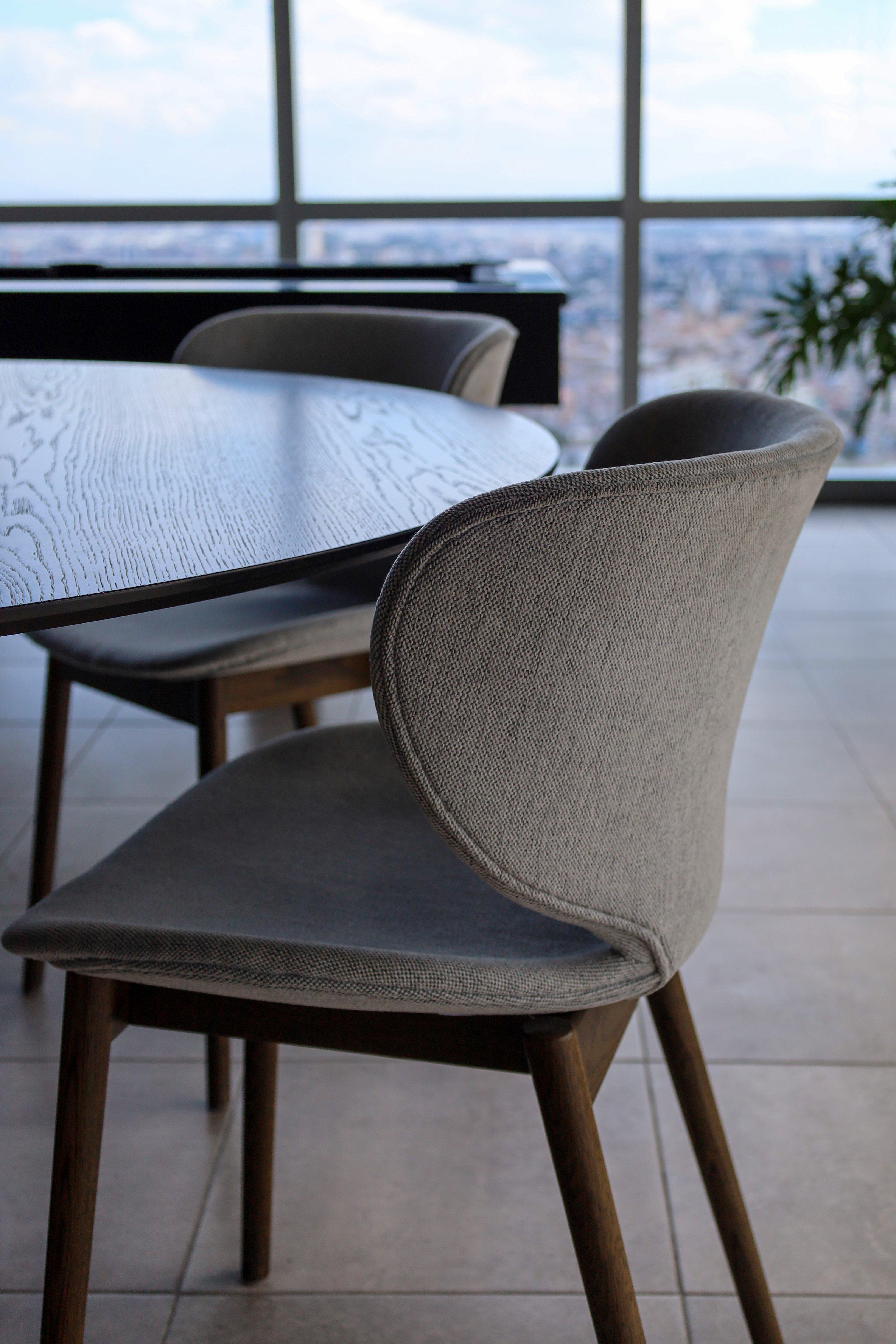 Grey upholstered chair beside a round wooden table in a room with large windows.