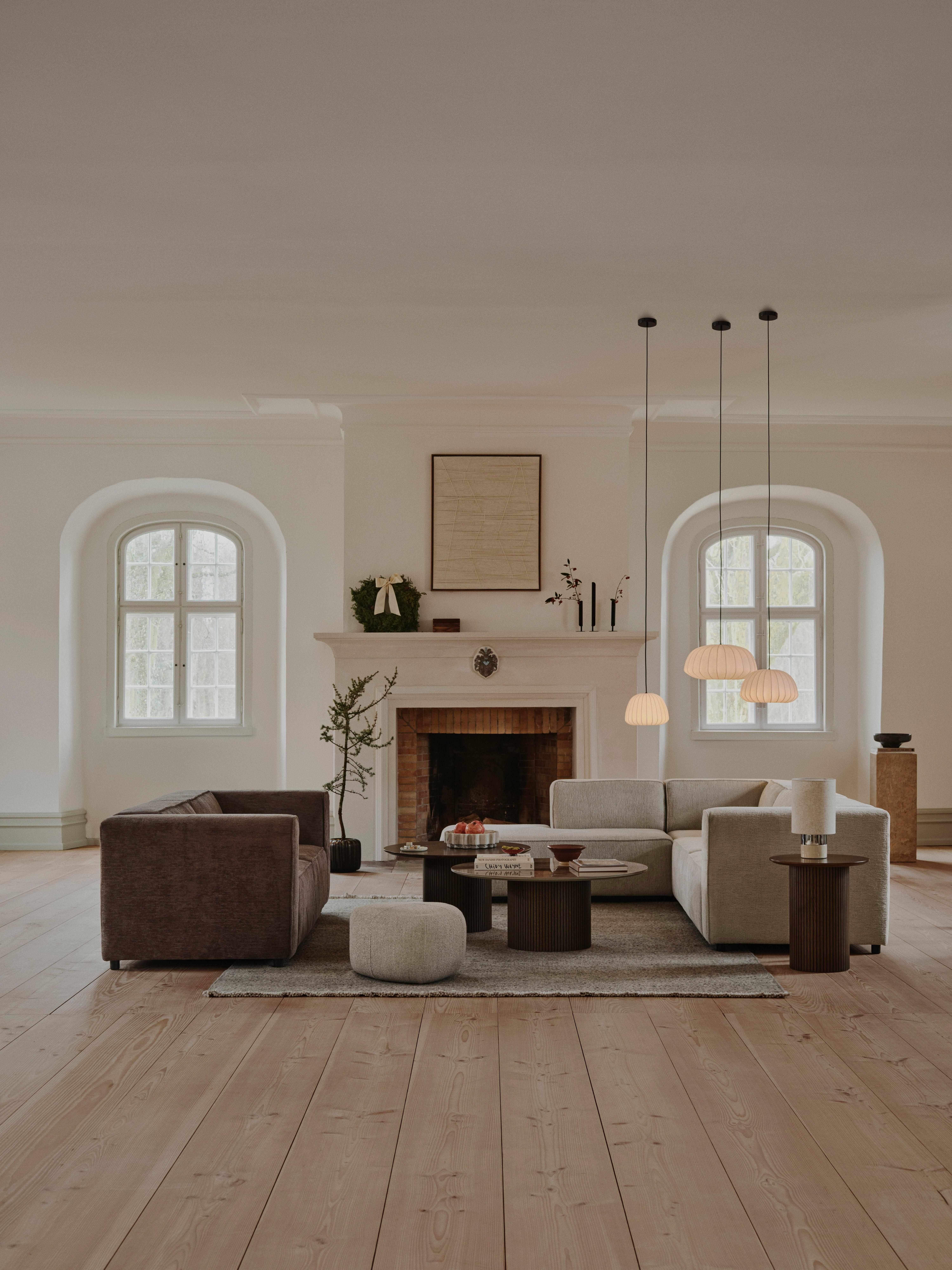 Spacious living room with large windows showcasing a forest view. The room features a Carmo corner sofa in camel Frisco fabric with Aquaclean, adorned with rusty orange velvet cushions, camel Frisco Salamanca cushions, caramel and beige Cord cushions, a cream Bouclé heavy cushion, and a pumpkin spice Bouclé cushion. The seating area includes a mustard Tuscany fabric Eden footstool and a golden beige Avellino fabric Bermuda footstool. A natural oak veneer oval Santiago coffee table sits on a beige and rusty orange Moments rug. Decor elements include a black and sand-colored Eternity Gallery wall art piece, a natural-colored Keep Me Warm plaid, and a warm grey Parasol bowl. Light wood flooring and soft natural light create a warm and inviting atmosphere.