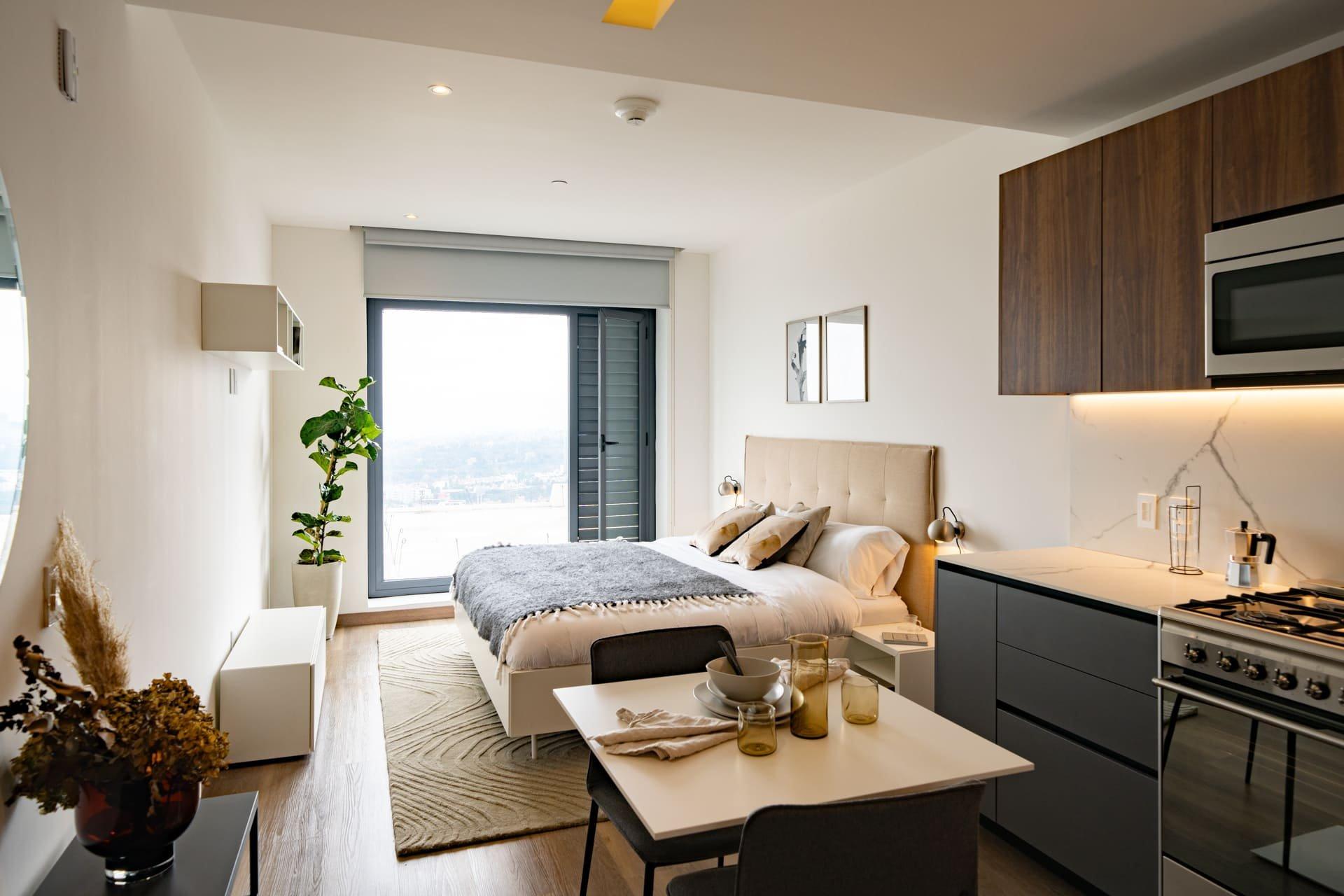 Studio apartment with bed, kitchenette, and dining area, natural light from window.