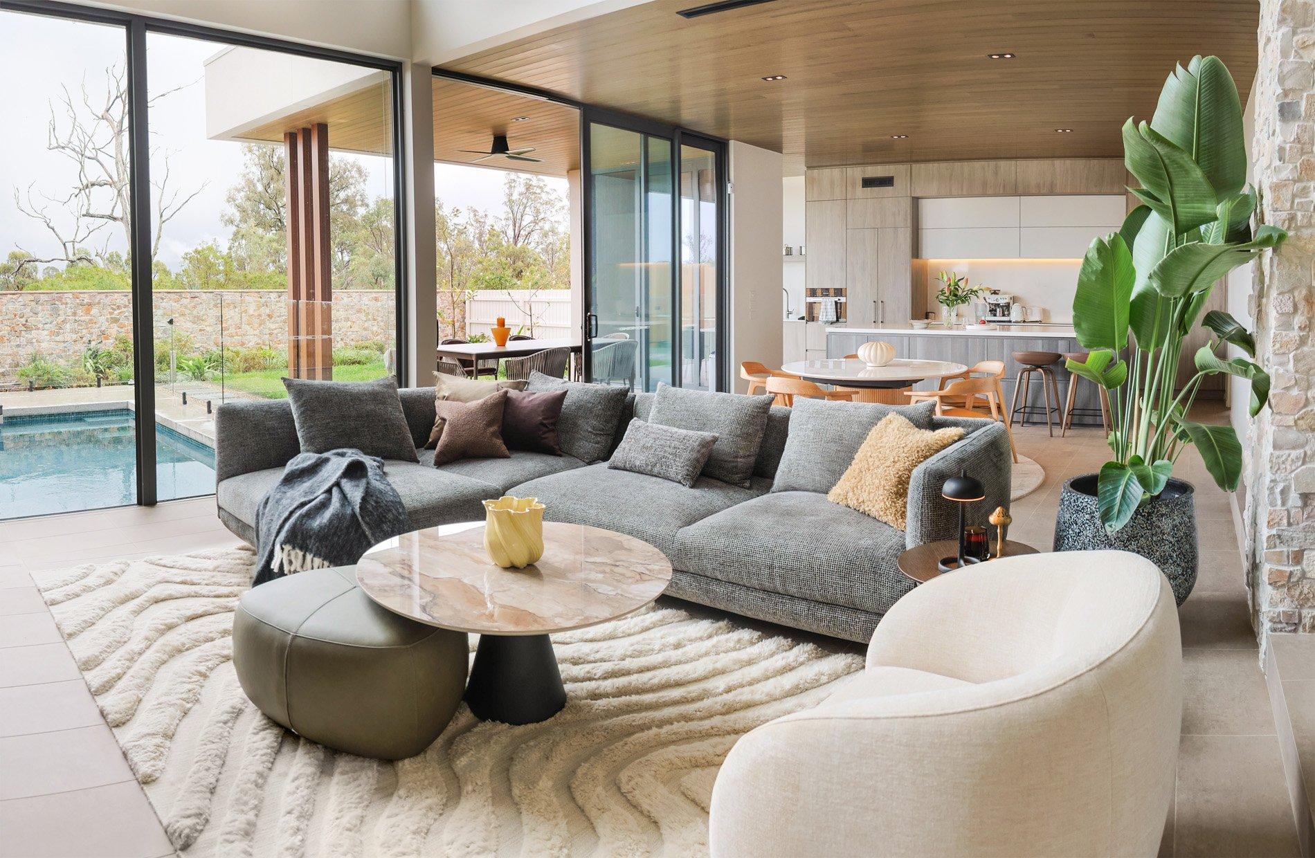 Moderne stue med grå sofa, beige stol, stort vindu mot basseng, og frodig plante.