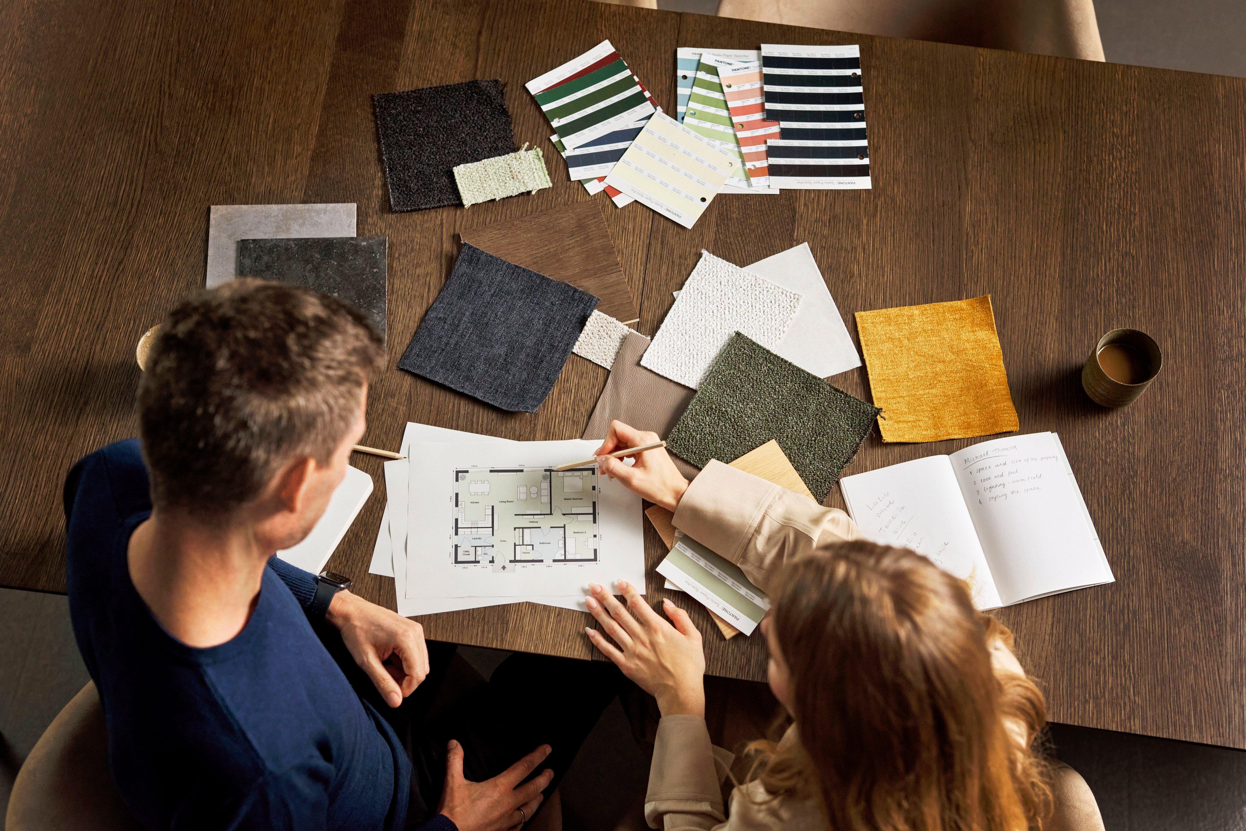 Two individuals reviewing a floor plan amid various fabric swatches on a wooden table.