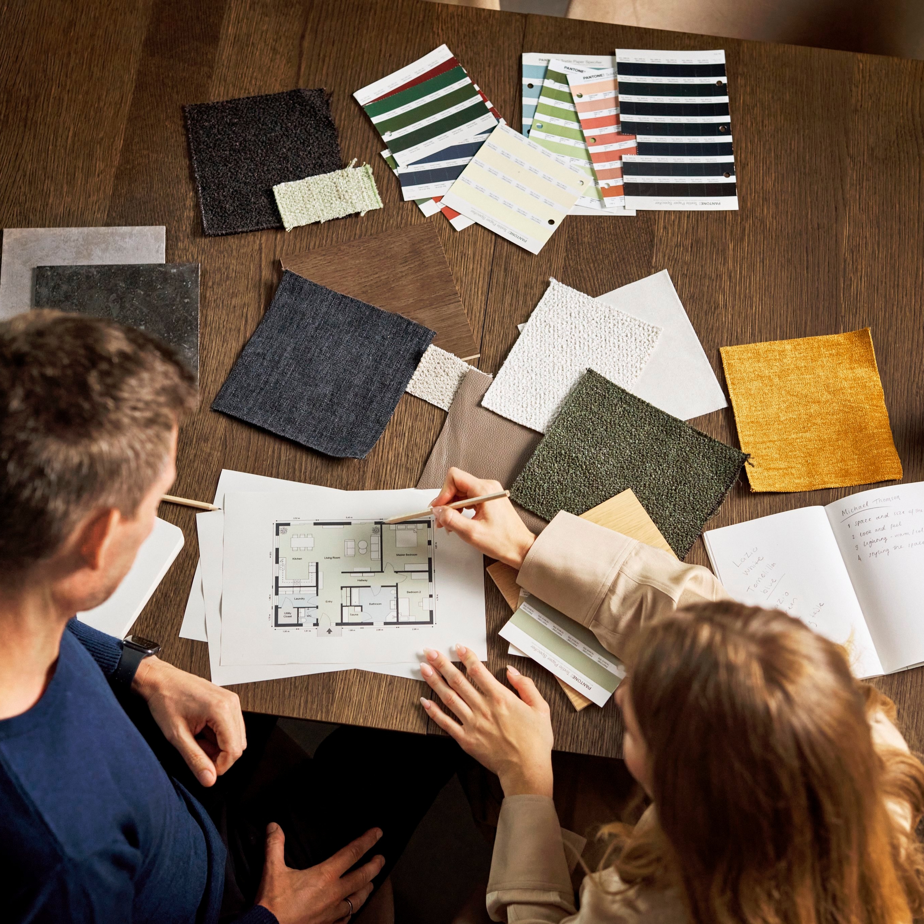 Two individuals reviewing a floor plan amid various fabric swatches on a wooden table.
