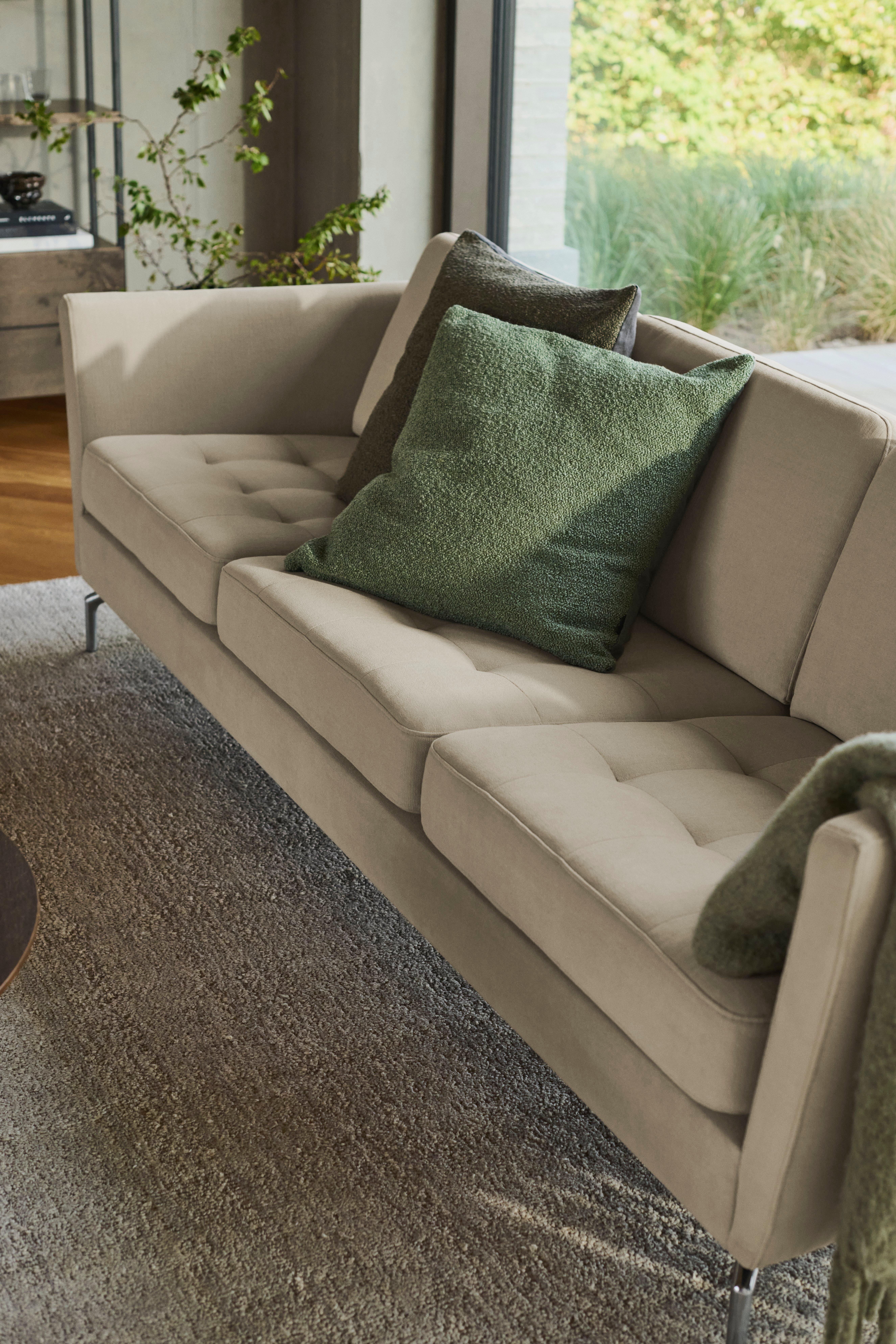 Close-up of a beige tufted Osaka sofa with green textured cushions and a throw blanket, placed on a gray rug near a window with greenery outside