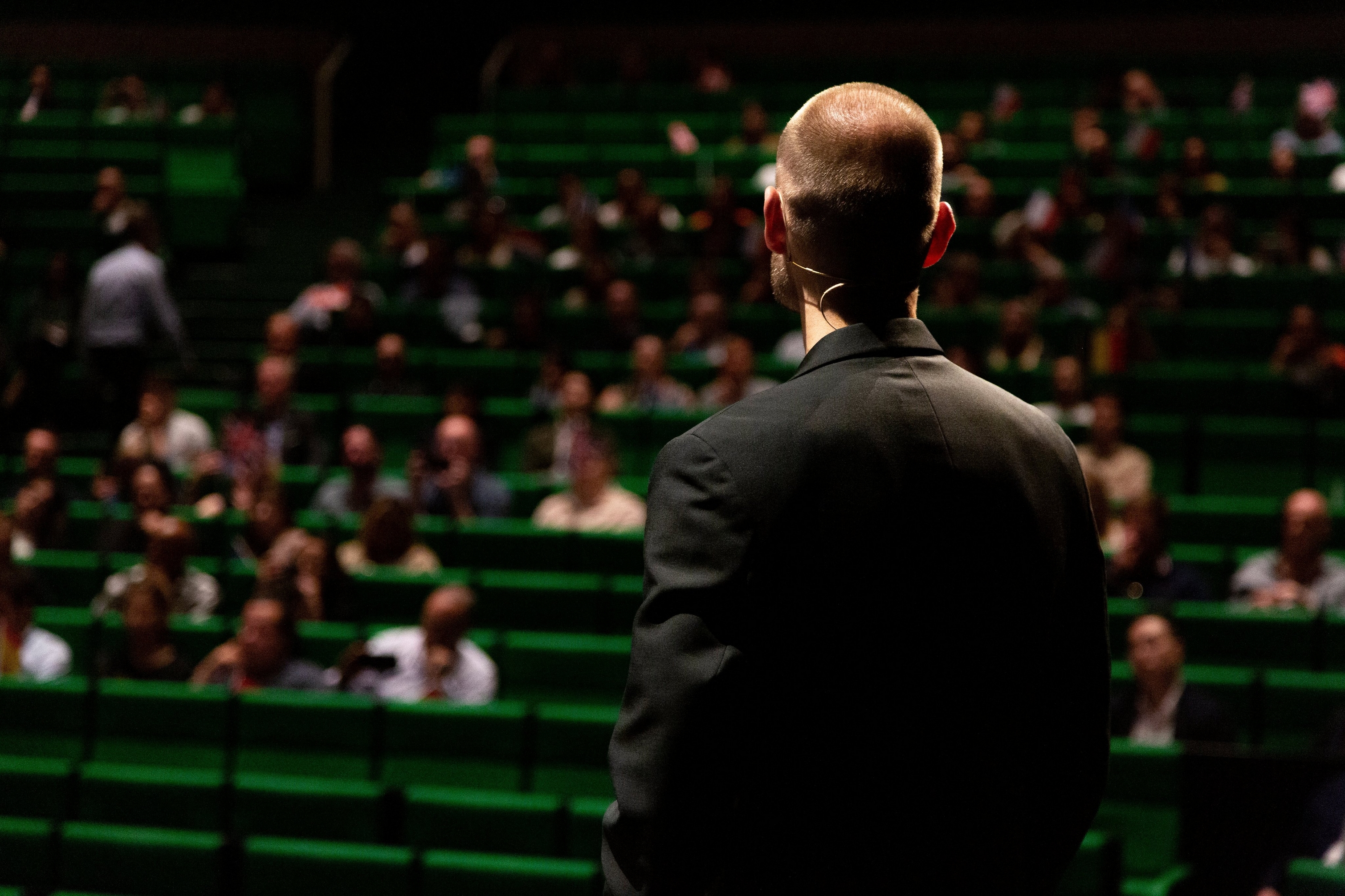 Hombre con traje negro frente a un público en un auditorio con asientos verdes.