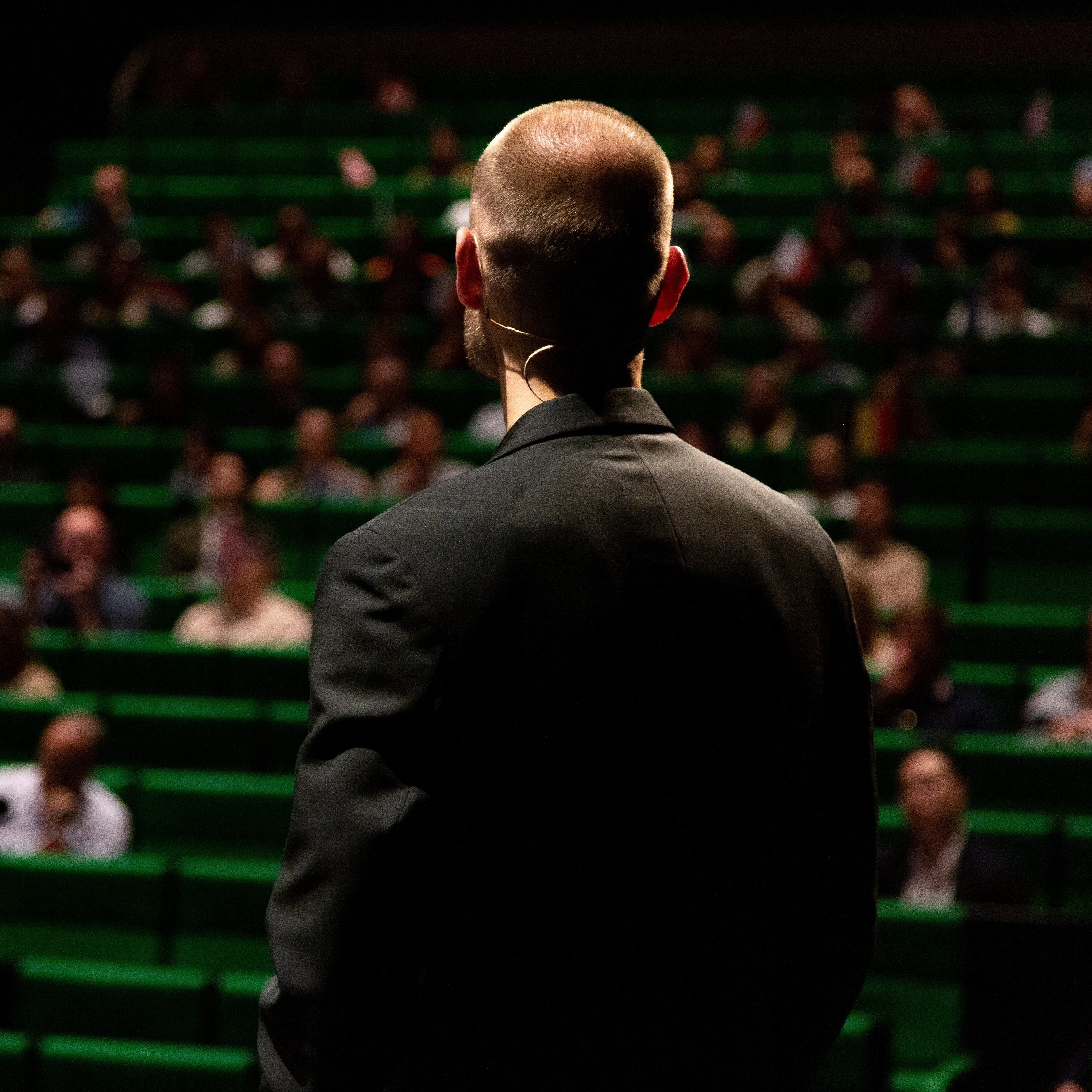 Man in black suit facing an audience in a green-seated auditorium.