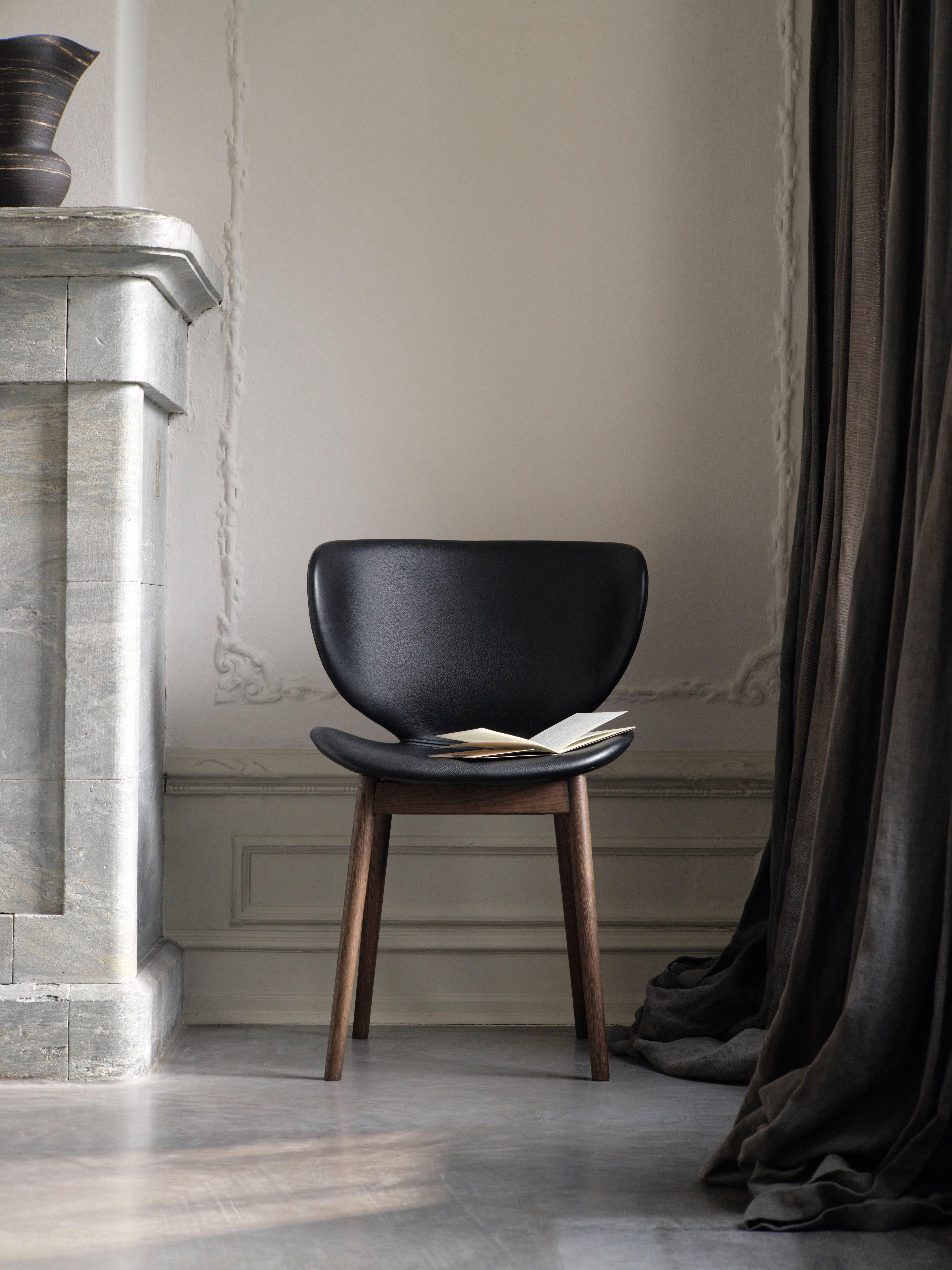 Black chair with curved seat and dark wooden legs, placed beside a stone mantel and long dark curtains on a gray floor.