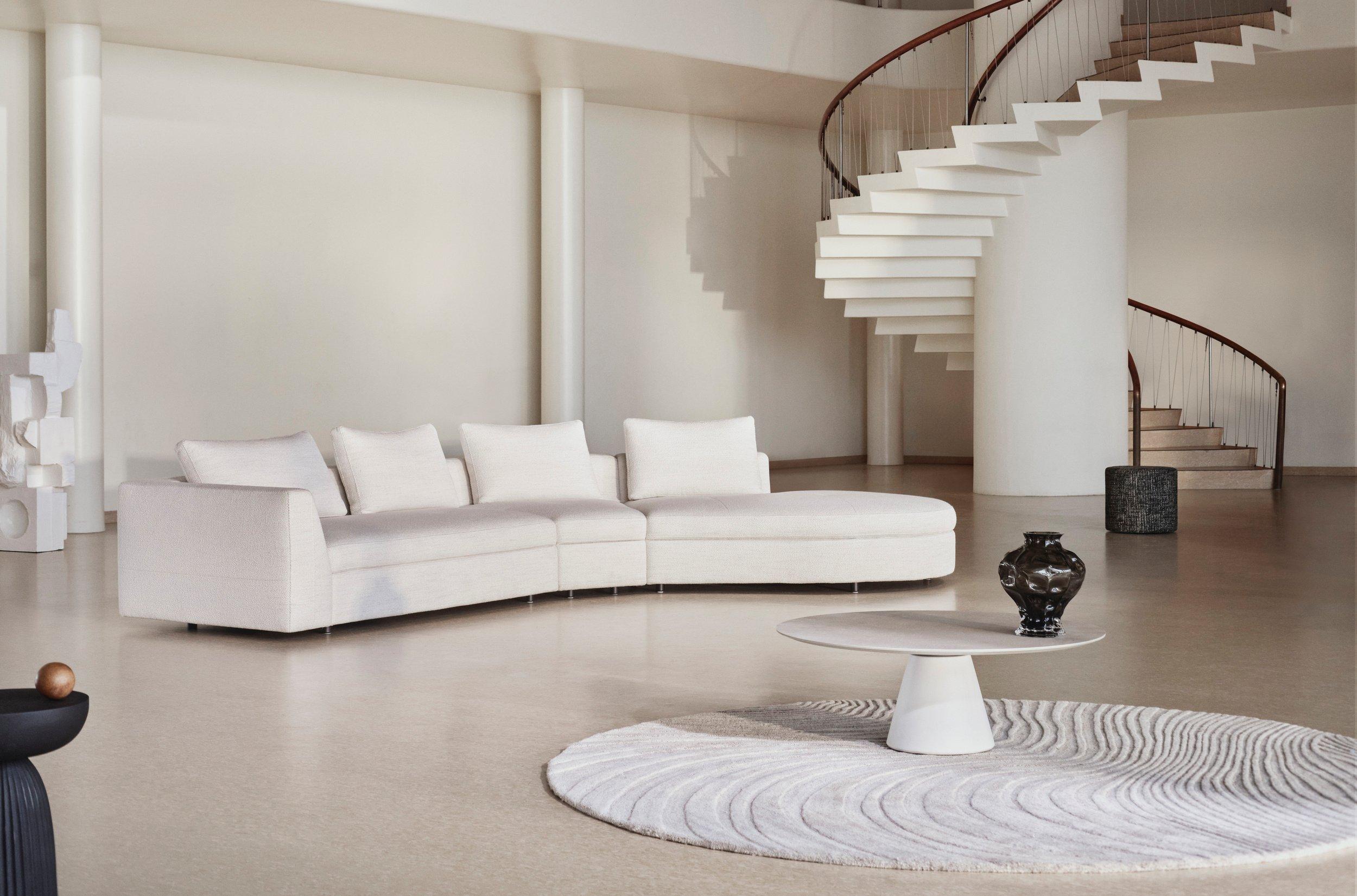 Divisão espaçosa com um sofá seccional branco curvo, escadaria em espiral escultural, tapete redondo e teto alto com padrão de tijolo arqueado.