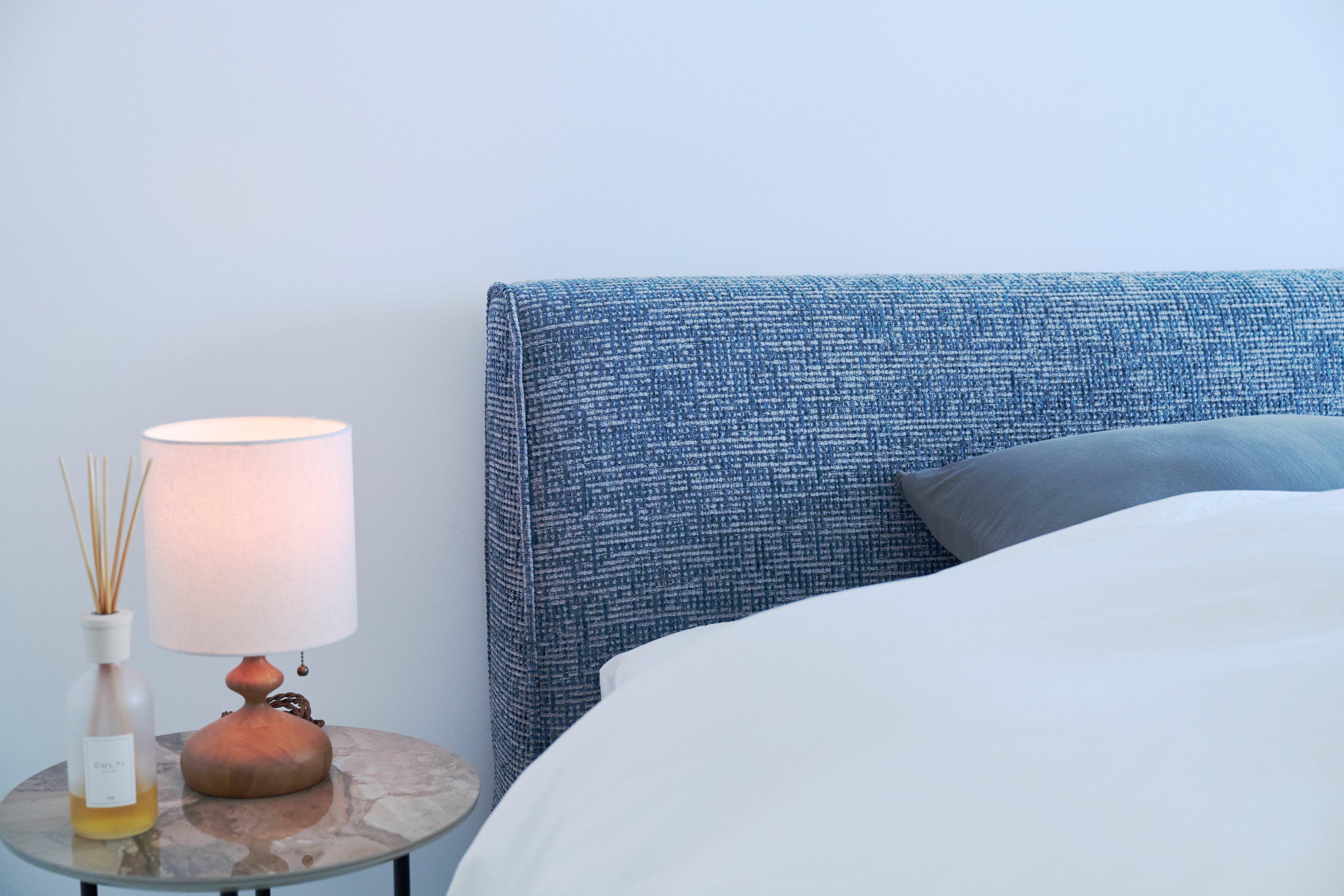 Blue upholstered headboard with bed linen next to a bedside lamp and reed diffuser.