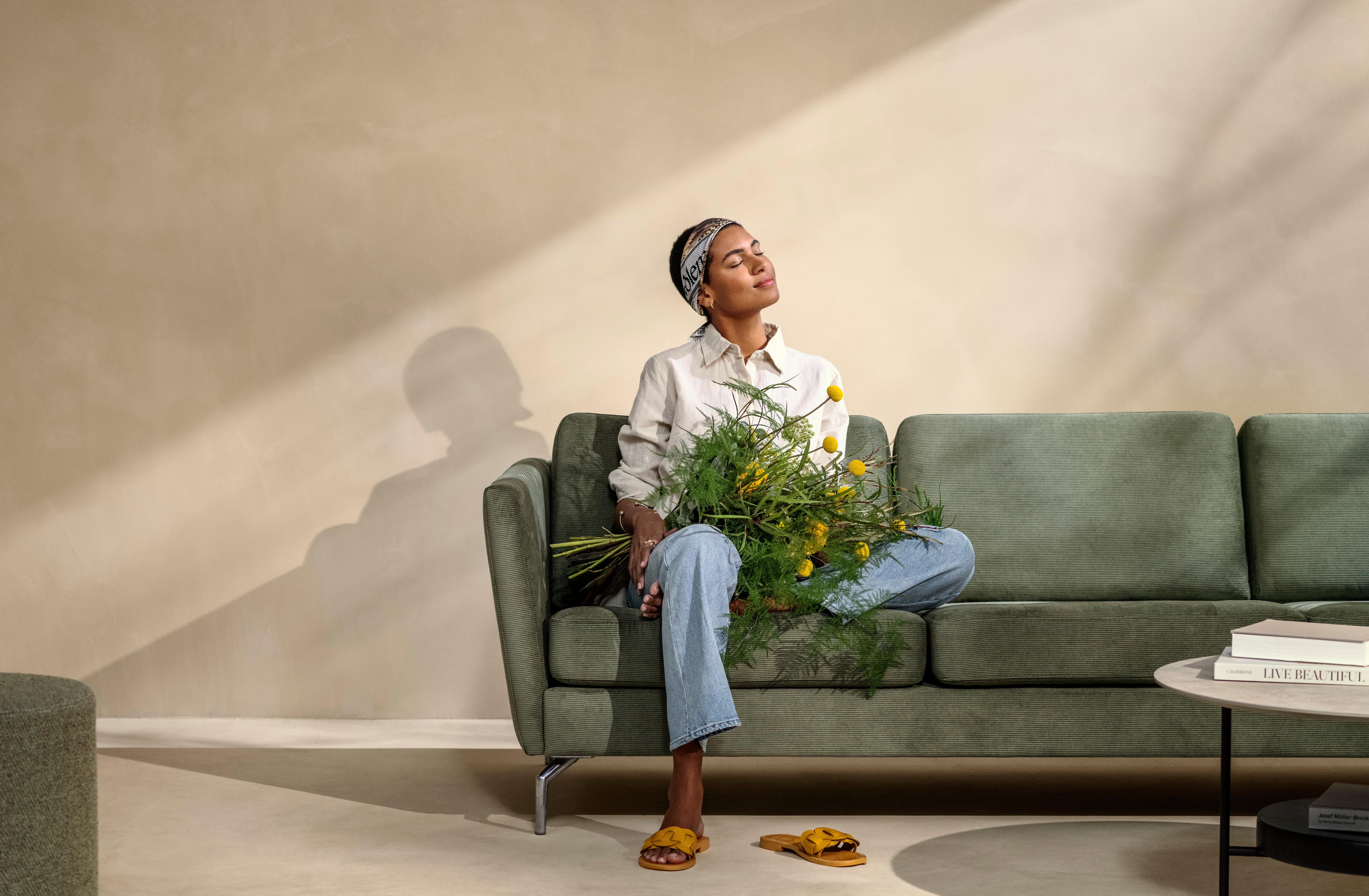 Woman holding flowers and sitting on the Osaka sofa in green Skagen fabric.