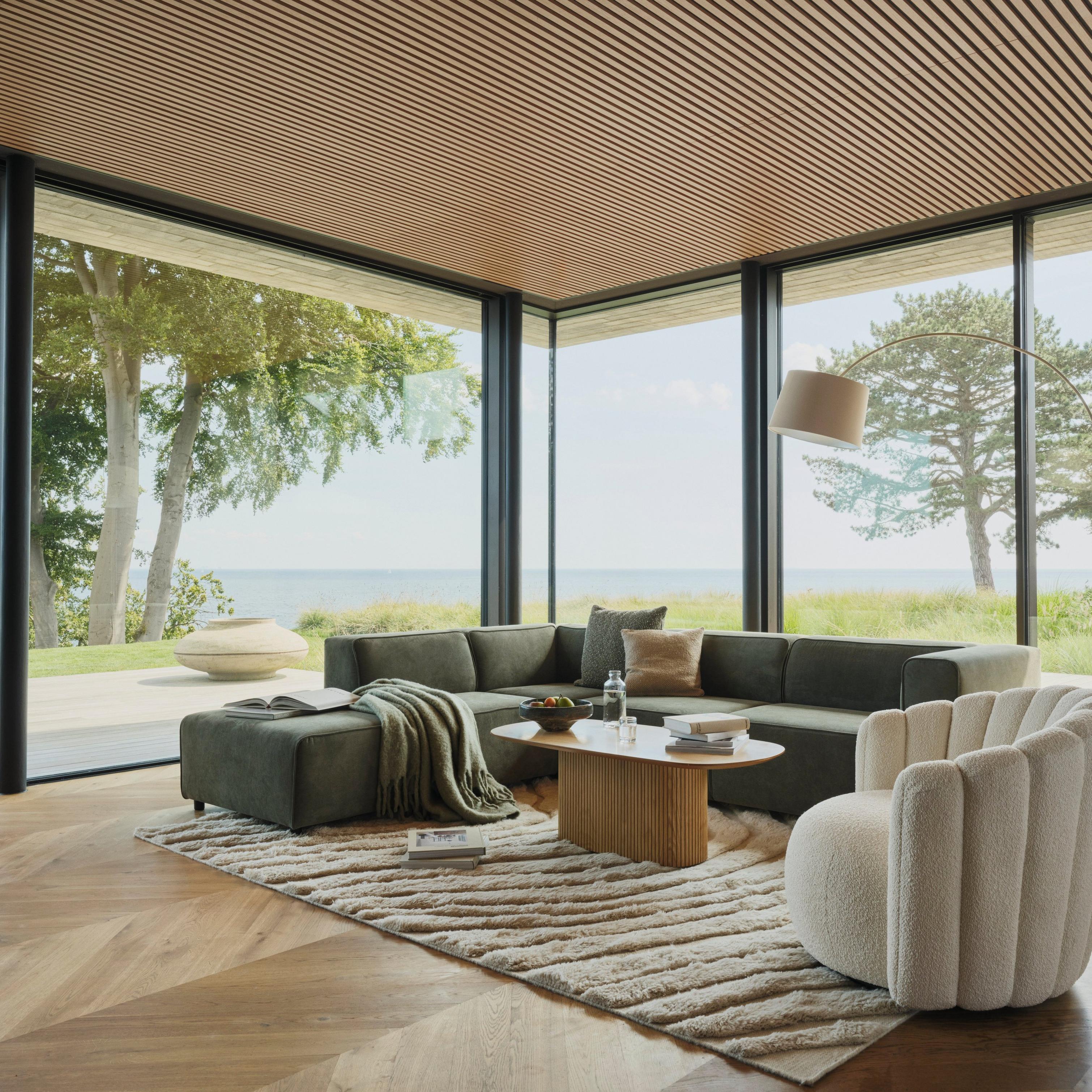 Modern living room with large windows, green sofa, cream chair, and wooden coffee table.