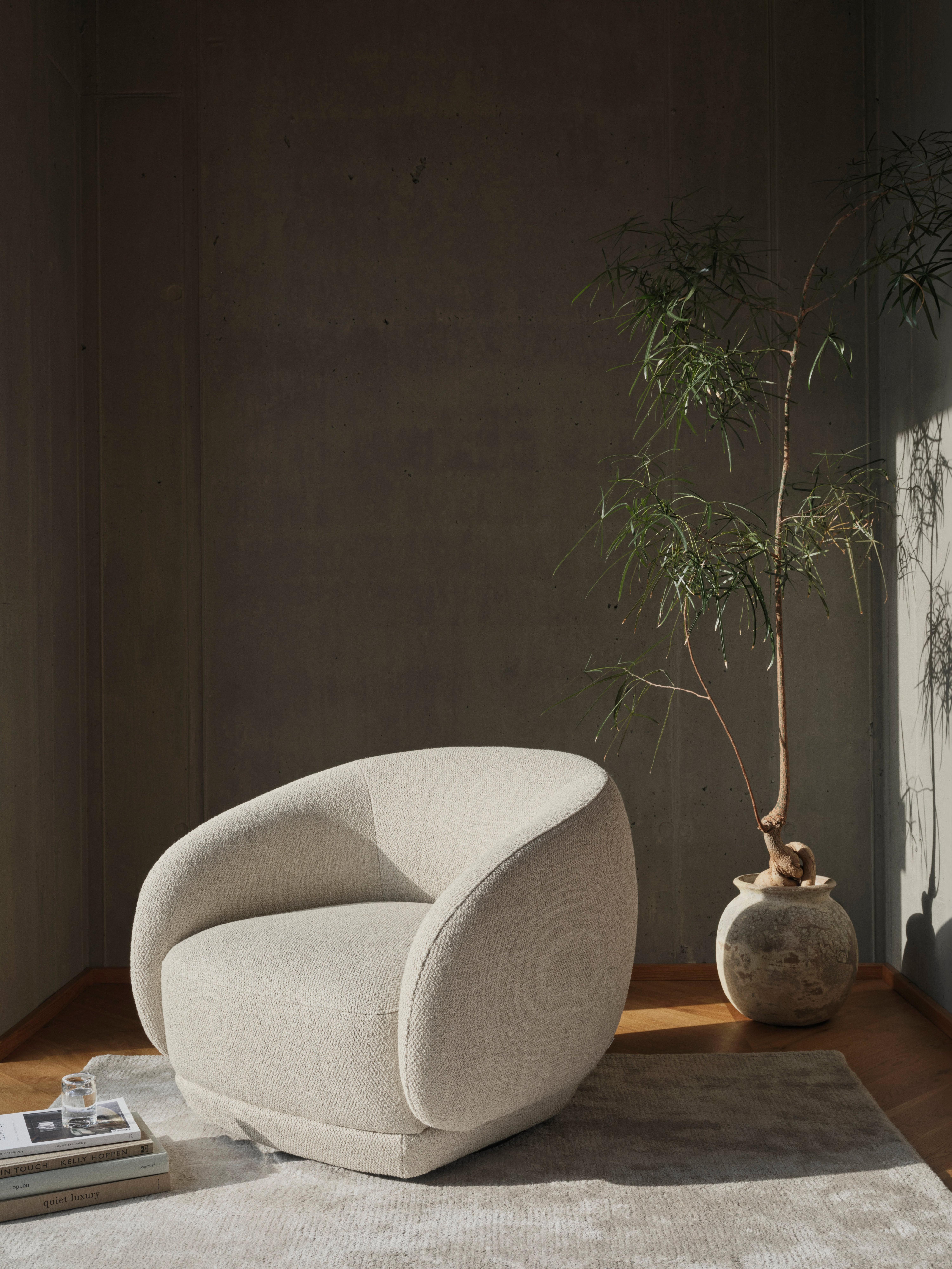Curved beige Bolzano armchair on light rug beside tall potted plant in minimalist room with dark wall and wooden floor.