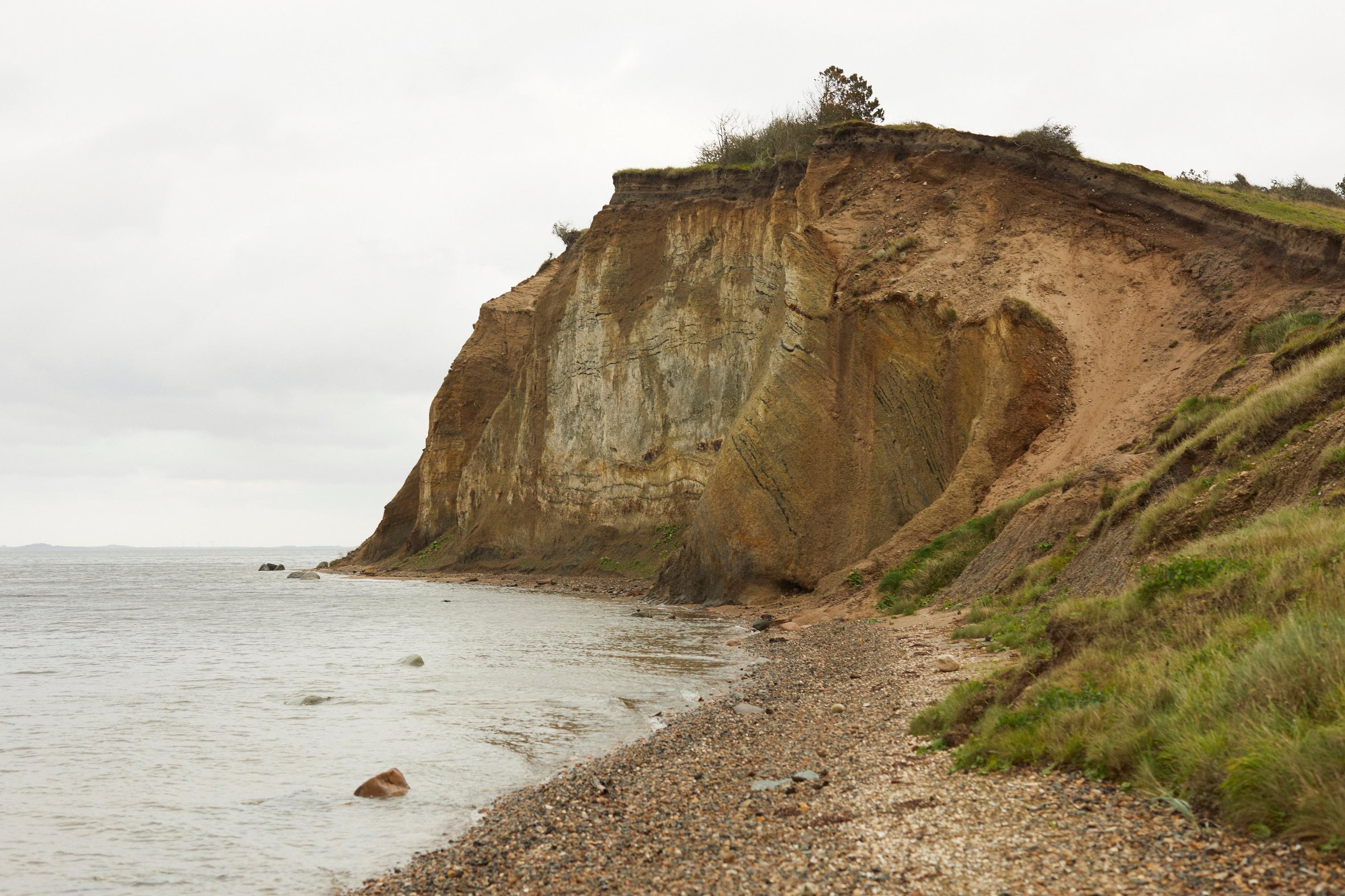 Cliff eroding into a pebble beach with calm sea under overcast sky.