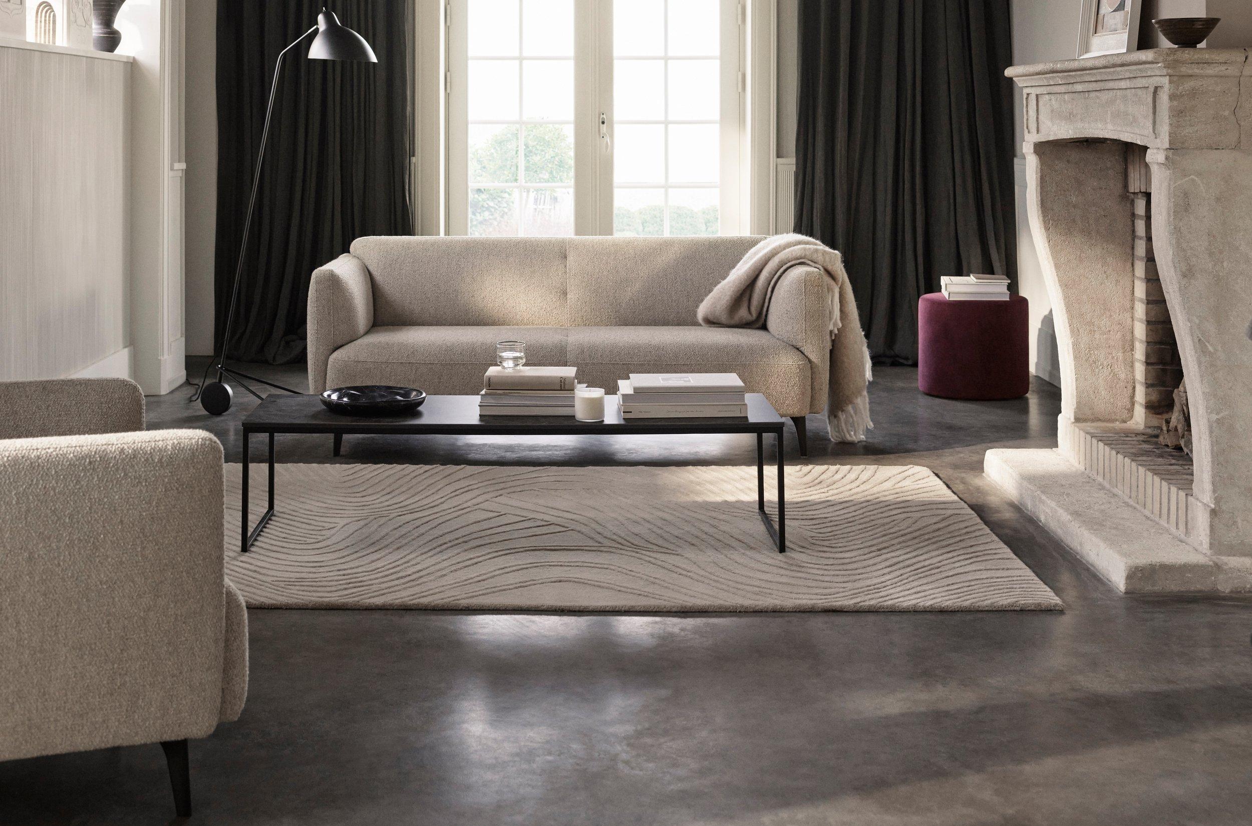 Modern living room with beige Modena sofa, a round coffee table on a striped rug, floor-to-ceiling windows, and a view of a lush outdoor landscape.