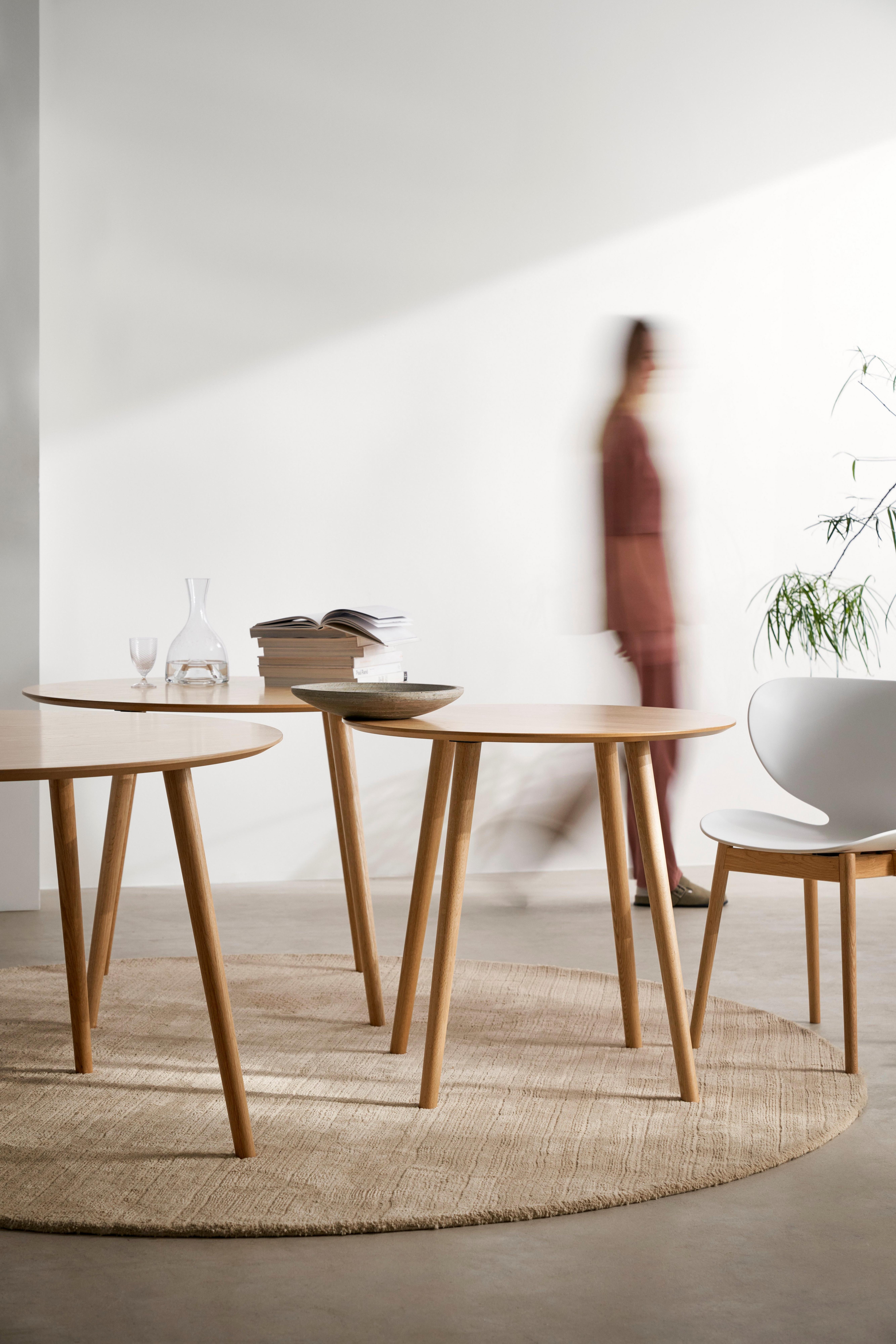 Three wooden Bornholm tables with a white Hamilton chair on a beige rug, with a glass decanter, books, and a bowl on the tables. A blurred person walks by in the background.