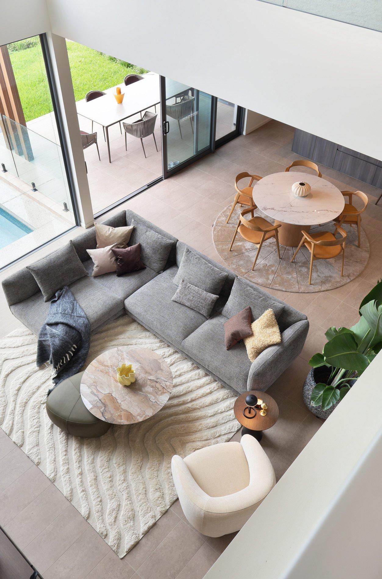 Grå hjørnesofa med puter, rundt marmorbord og lysebrun stol i stue. Rektangulært spisebord ute.