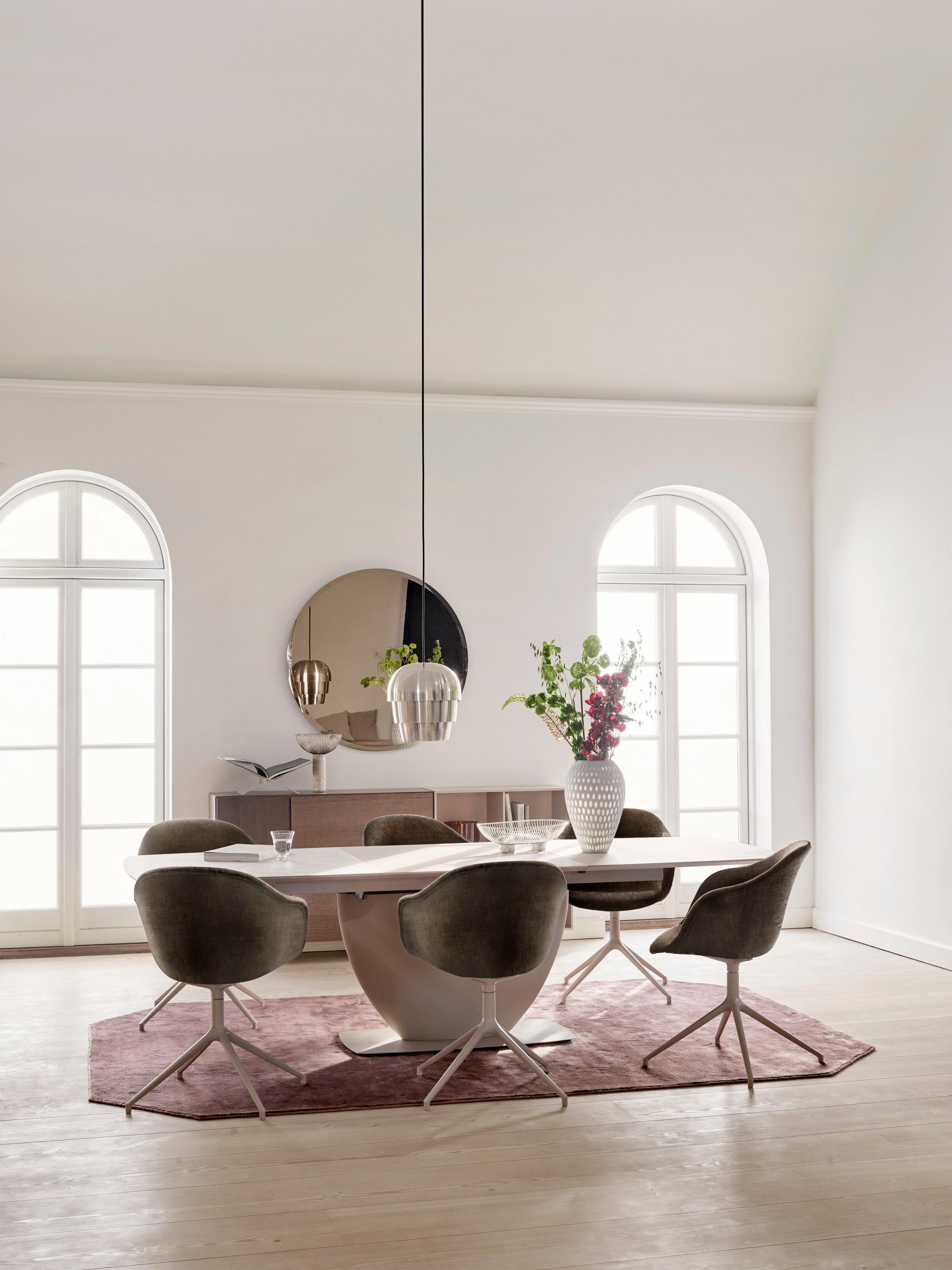 Elegant dining room with arched windows, a marble table, grey chairs, a round mirror, and floral accents.