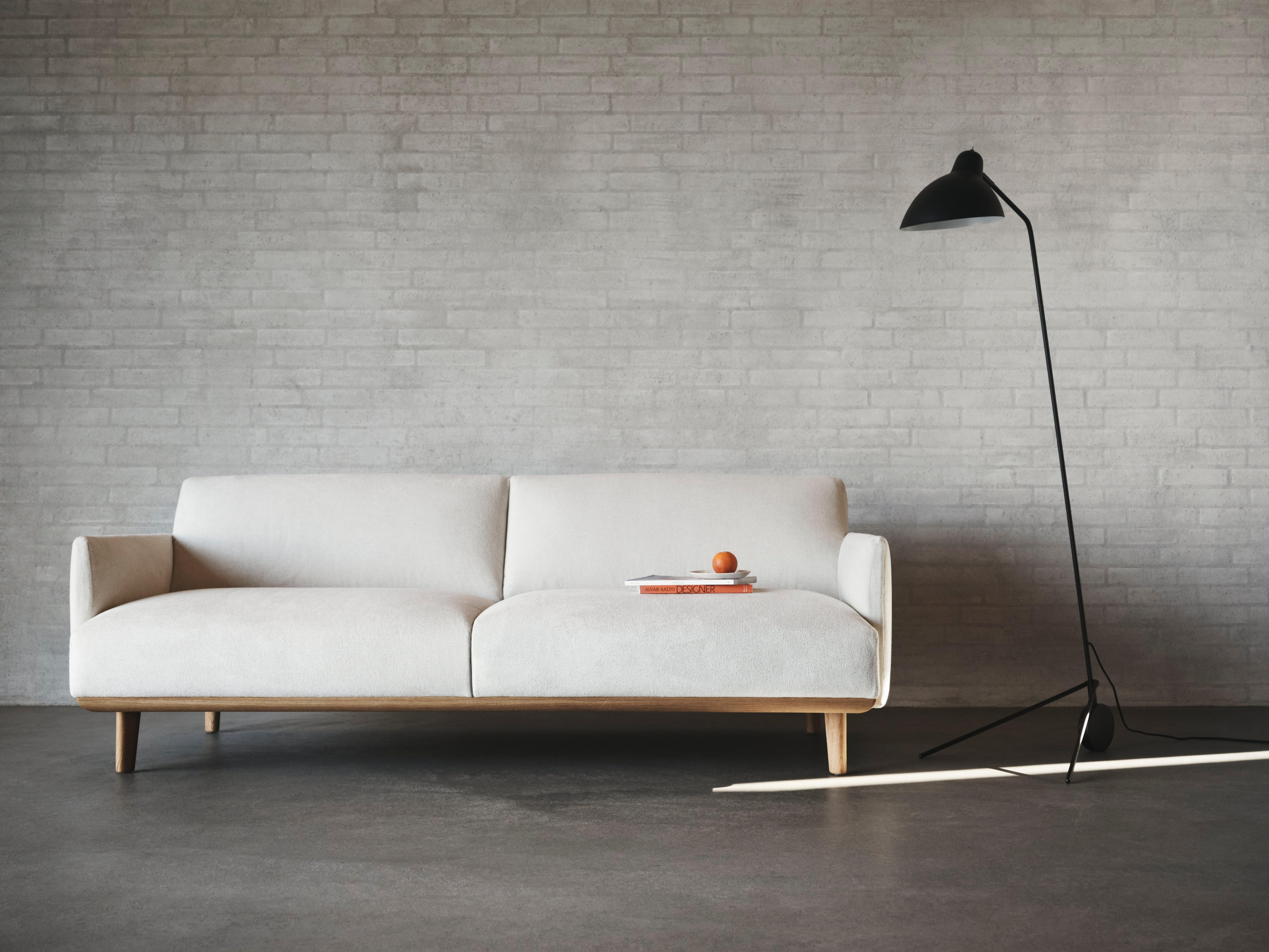 Minimalist white sofa with wooden base against a light grey brick wall, beside a black floor lamp
