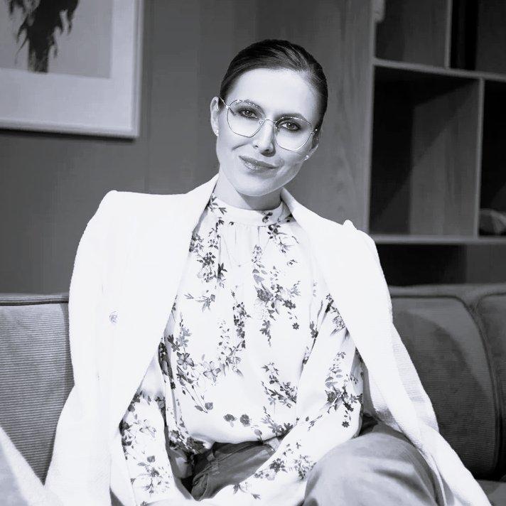 Woman wearing round glasses and a floral blouse with a blazer draped over her shoulders.