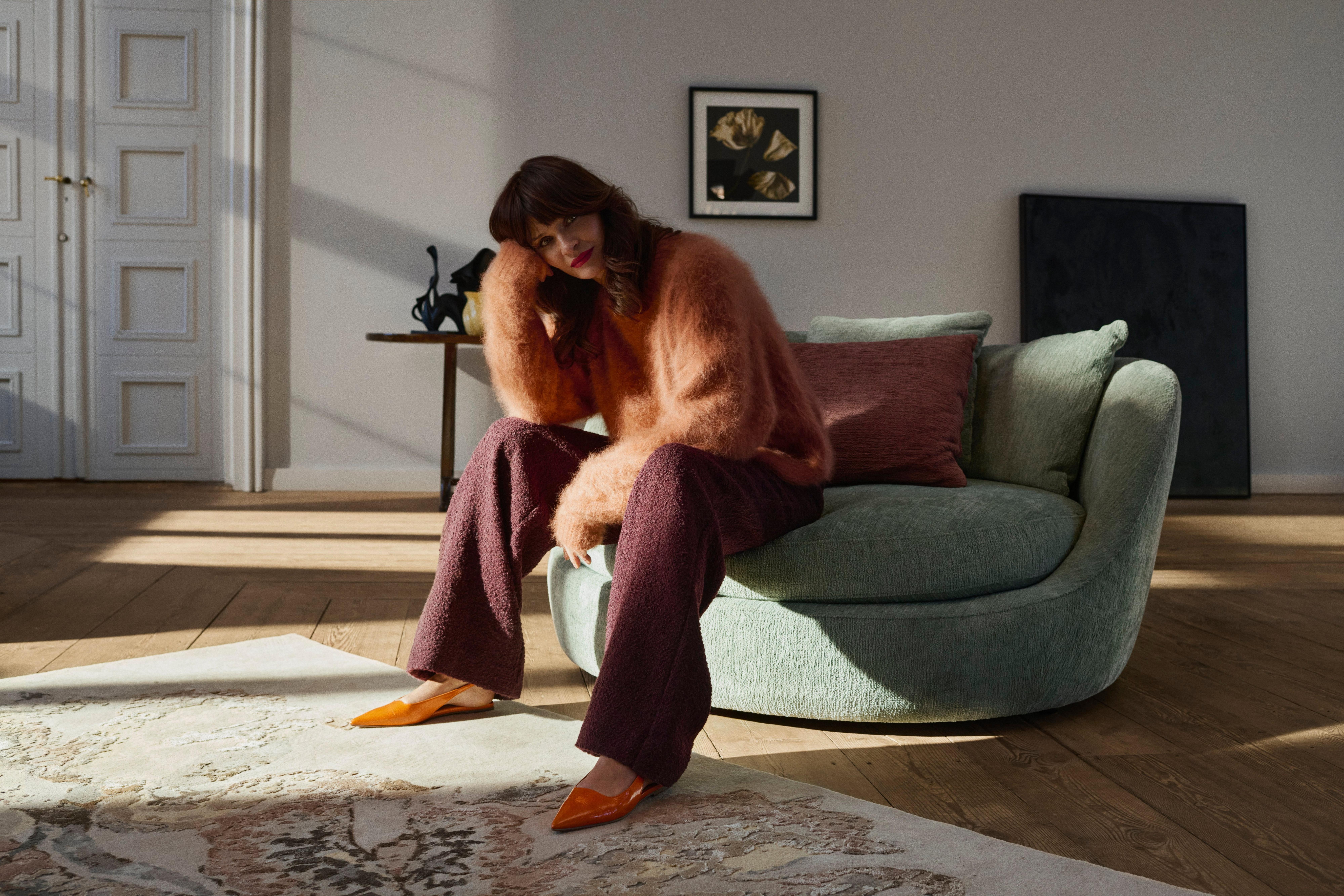 Helena Christensen seated on a sage green Ø lounge chair in a sunlit room with wood floors, artwork, and a light-toned patterned rug.