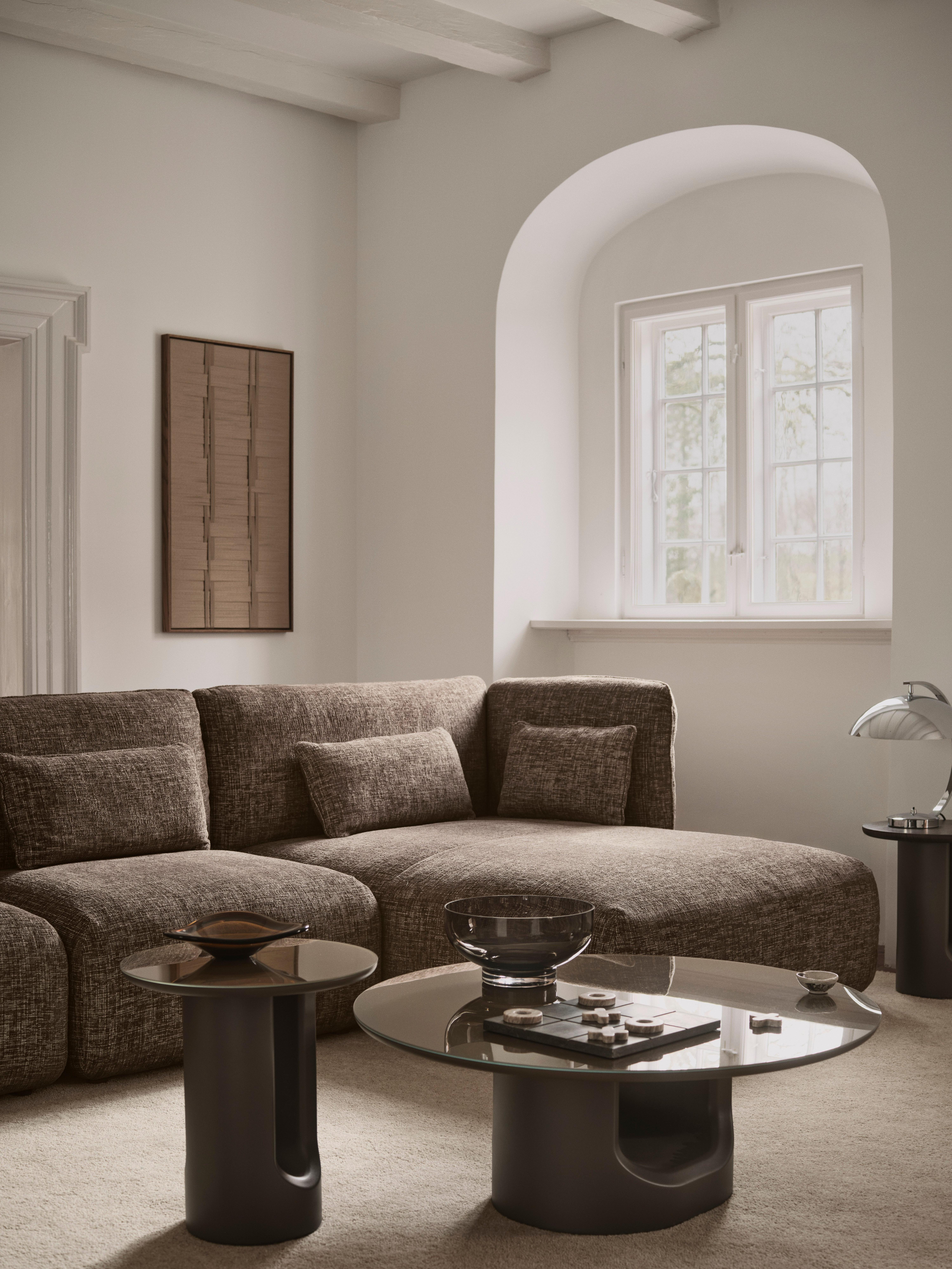 Brown fabric corner sofa with cushions, styled with round coffee tables and a table lamp.
