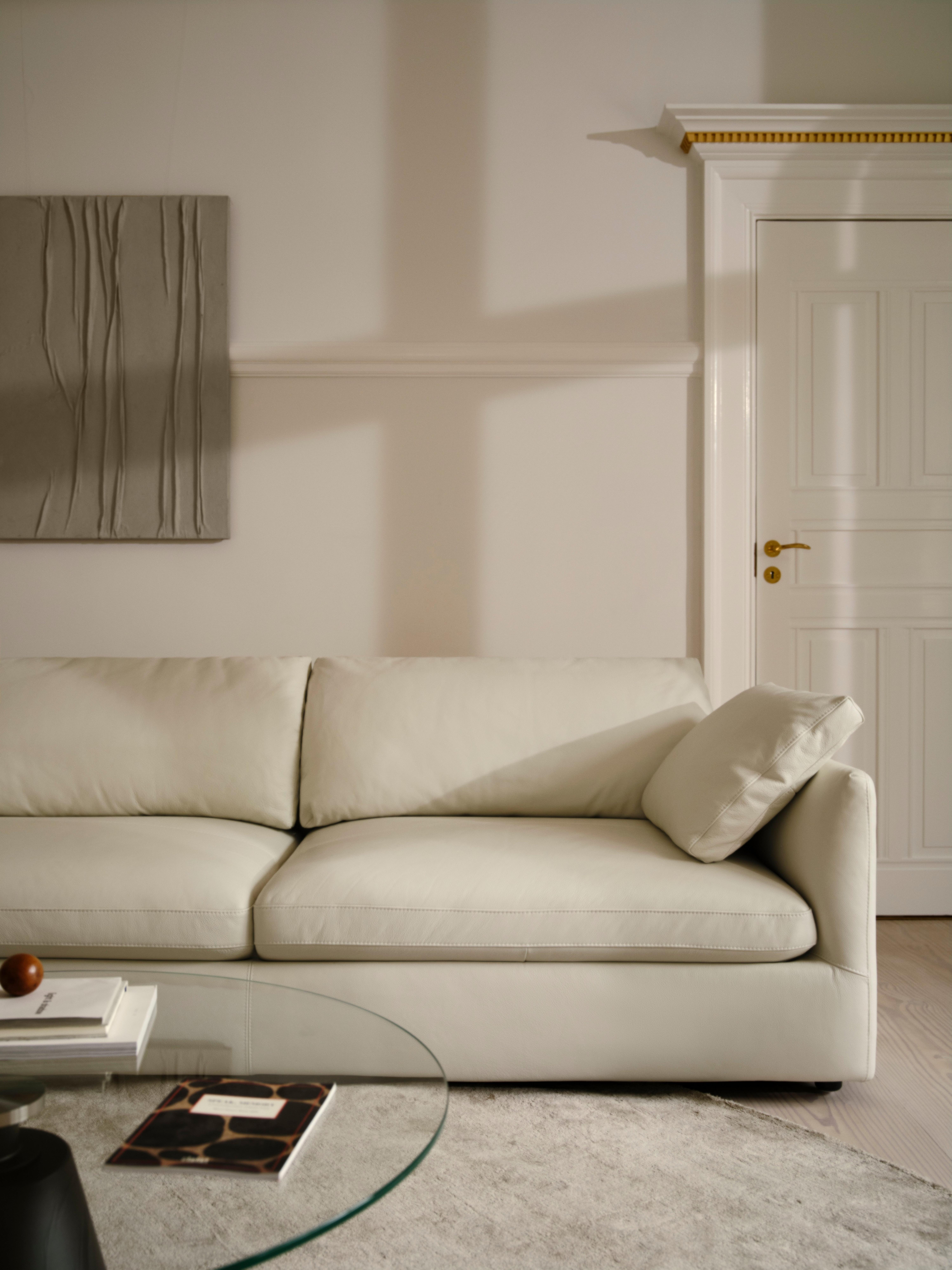 Sofá bege em sala de estar com mesa de vidro redonda e livros. Quadro abstrato na parede.