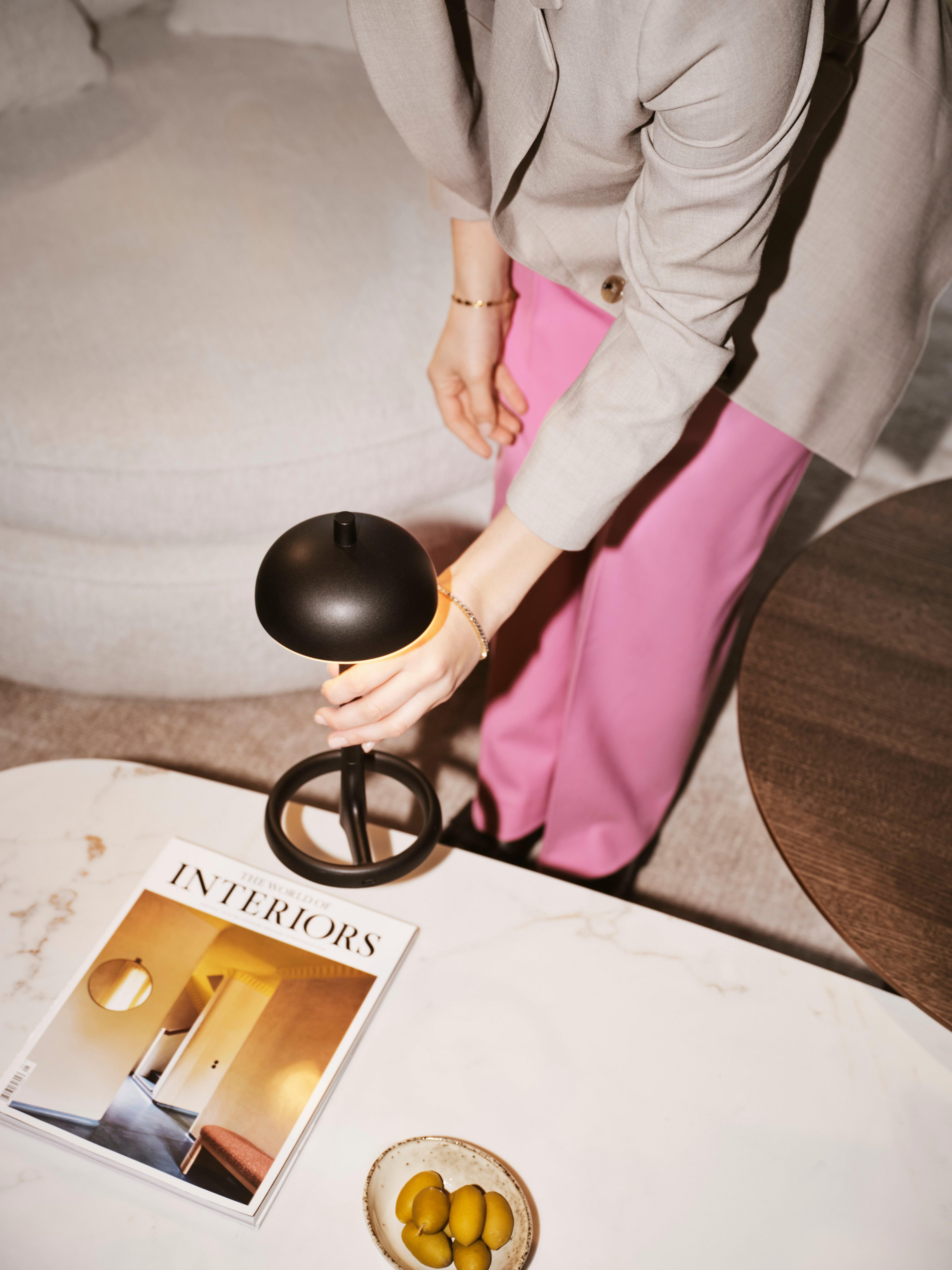 A matt black Paris table lamp sits on a Santiago oval coffee table with a white ceramic top, next to a bowl of olives and a magazine. To the side is the Santiago coffee table in dark oak veneer, and in the background, the Ø lounge chair in white Lucca fabric, adds a soft touch.