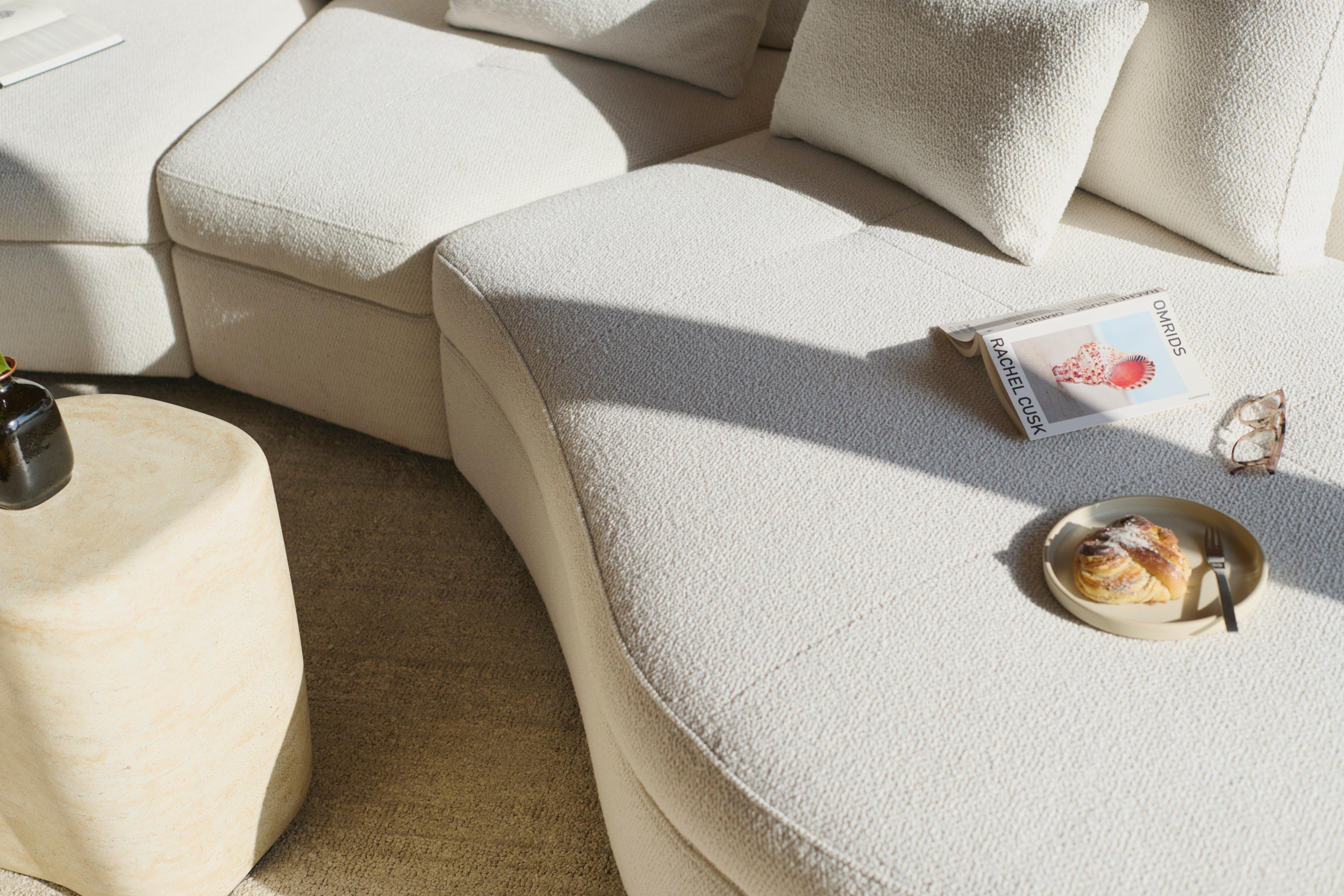 Image of the Bergamo sofa with round lounging unit in white Lazio fabric, with the Bergamo deco cushions in white Lazio fabric, the Terracina side table in Travertine look and the Serge rug light grey