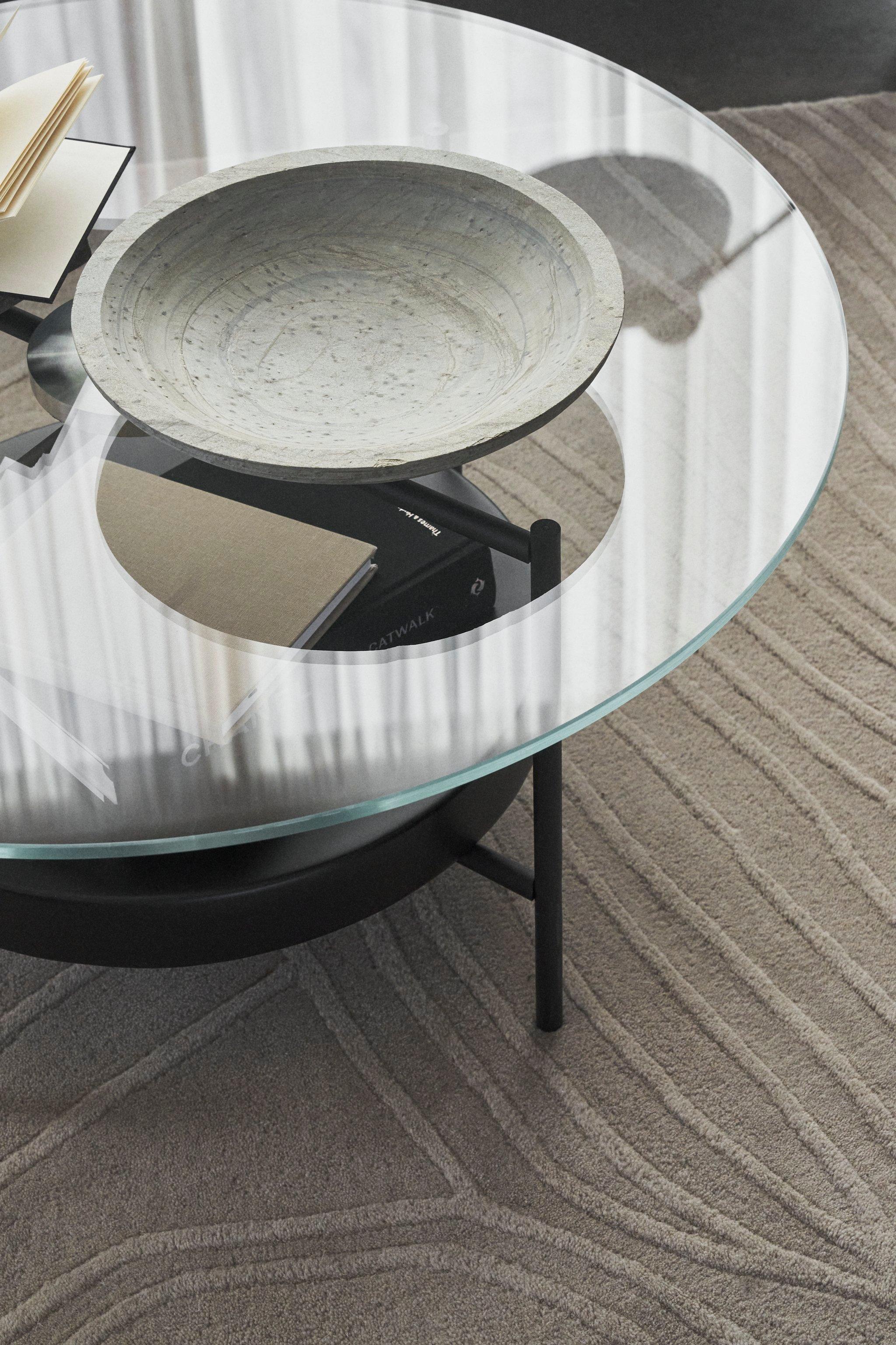 Moderne salontafel met glazen blad, een decoratieve schaal en boeken op een karpet met textuur.